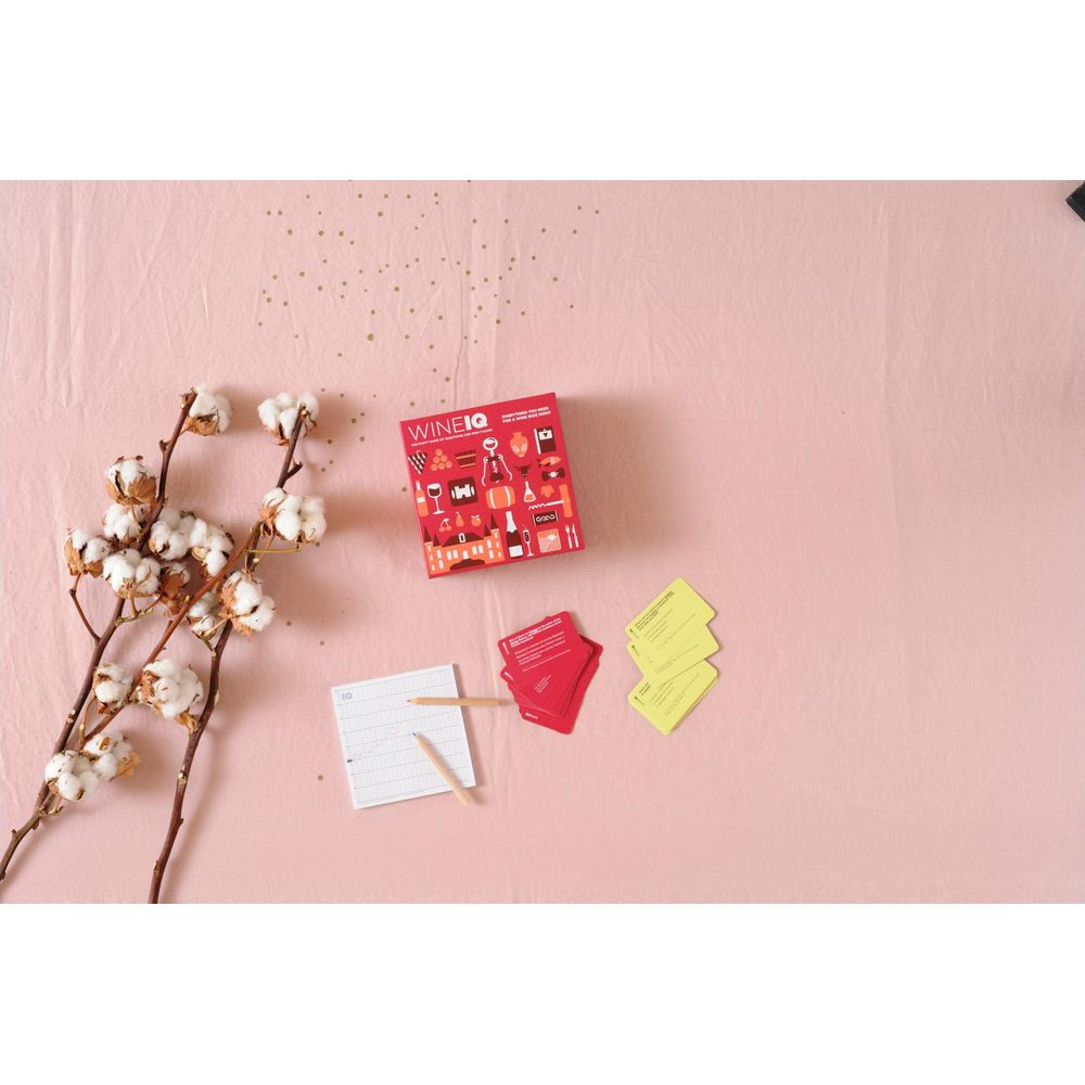 Une boîte rouge intitulée Wine IQ avec des cartes et des branches de coton sur une surface rose.