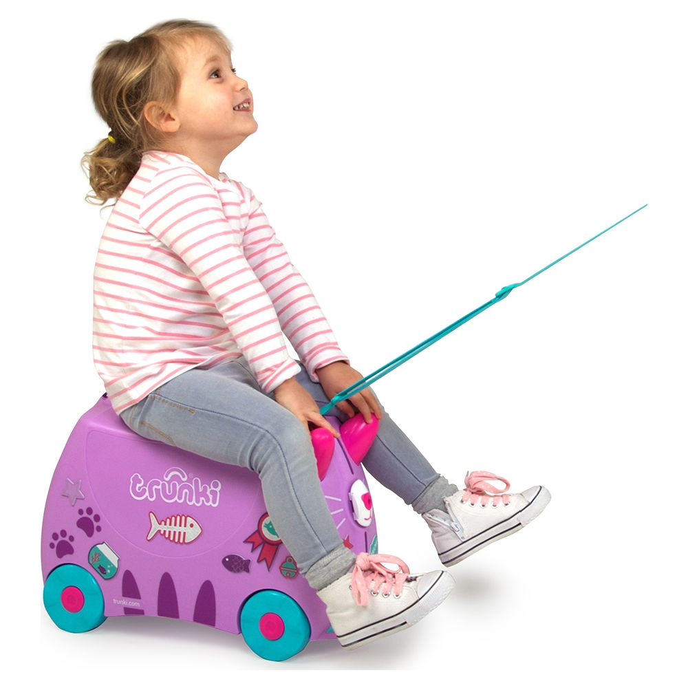 Una bambina è seduta su una valigia viola a forma di gatto, guardando in alto.