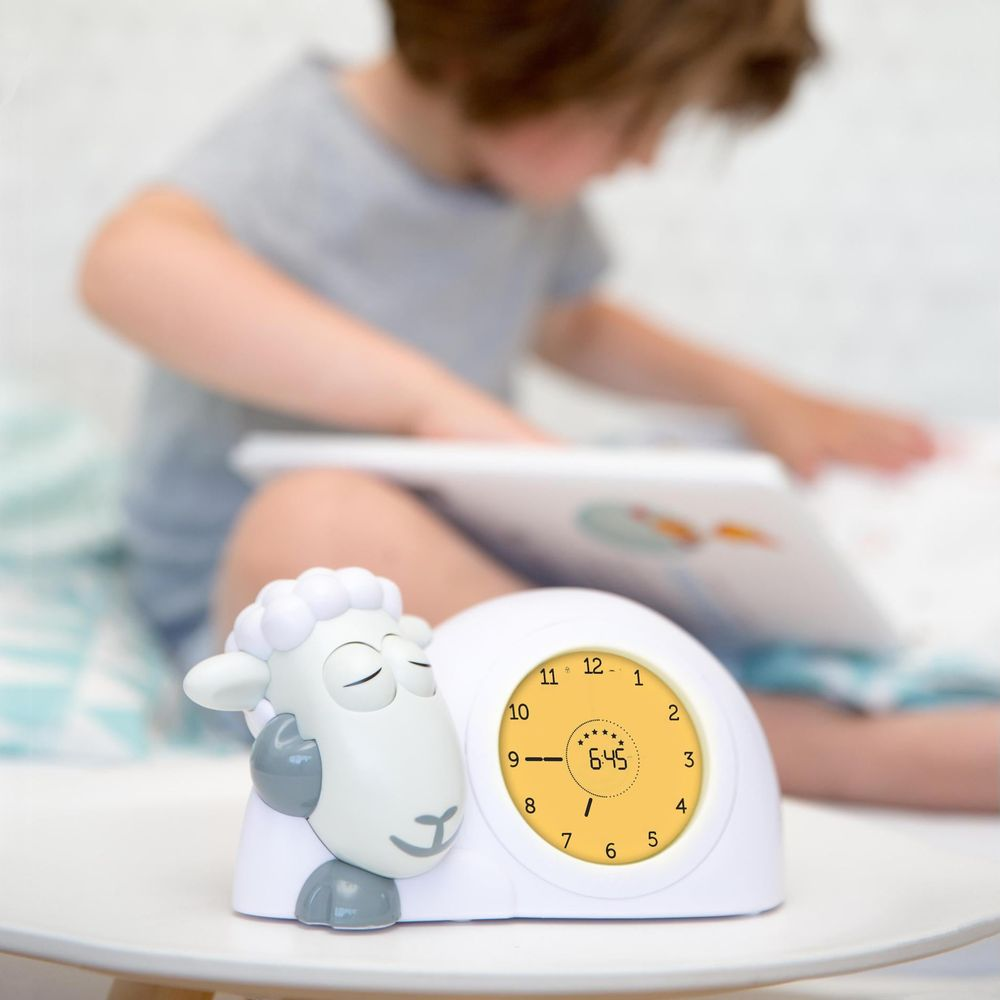 Ein Kinderwecker in Schafform zeigt 6:45 Uhr an, während ein Kind im Hintergrund liest.