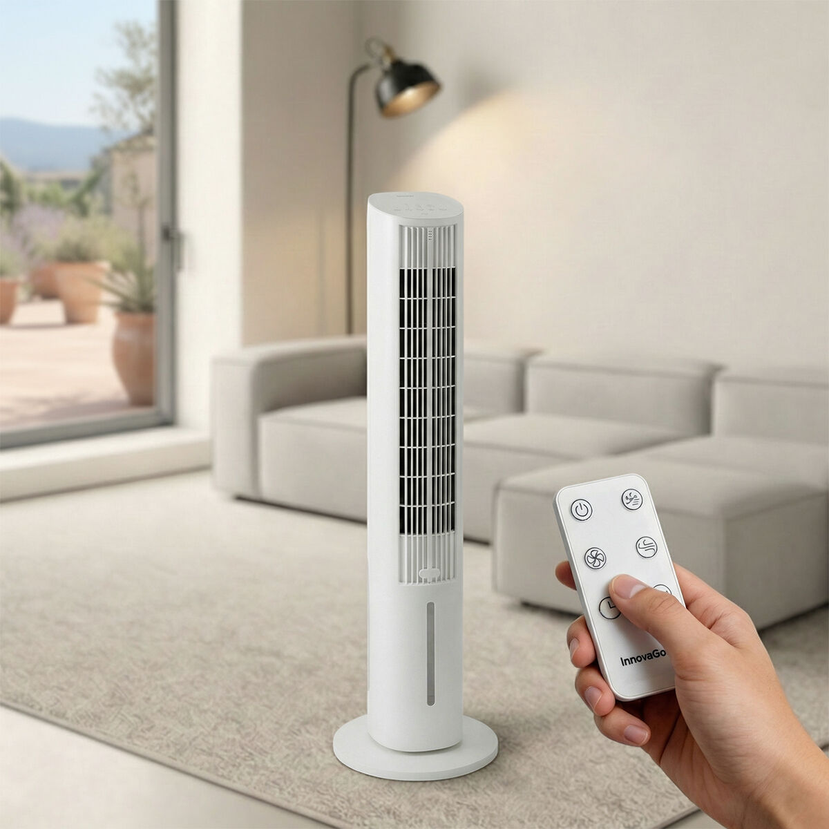 Un ventilador de torre blanco está sobre una alfombra en una sala, una mano sostiene un control remoto.