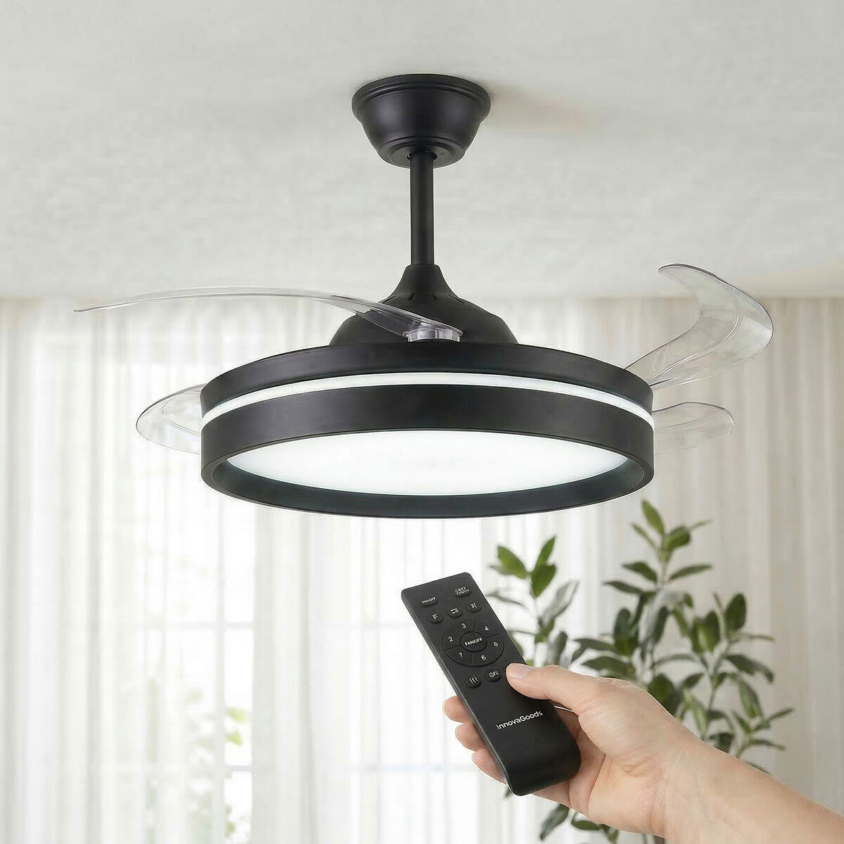Un ventilatore da soffitto nero con luce LED bianca e pale retrattili trasparenti, controllato da una mano che tiene un telecomando.