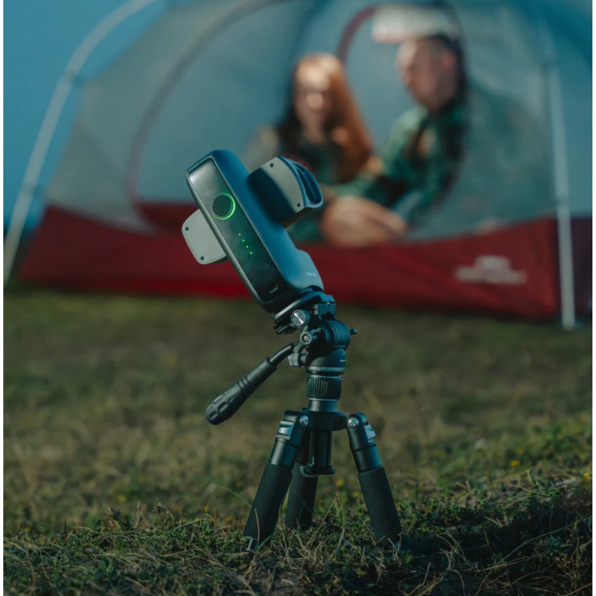 Ein schwarzer Gimbal mit grüner LED auf einem Stativ vor einem Zelt mit Personen.