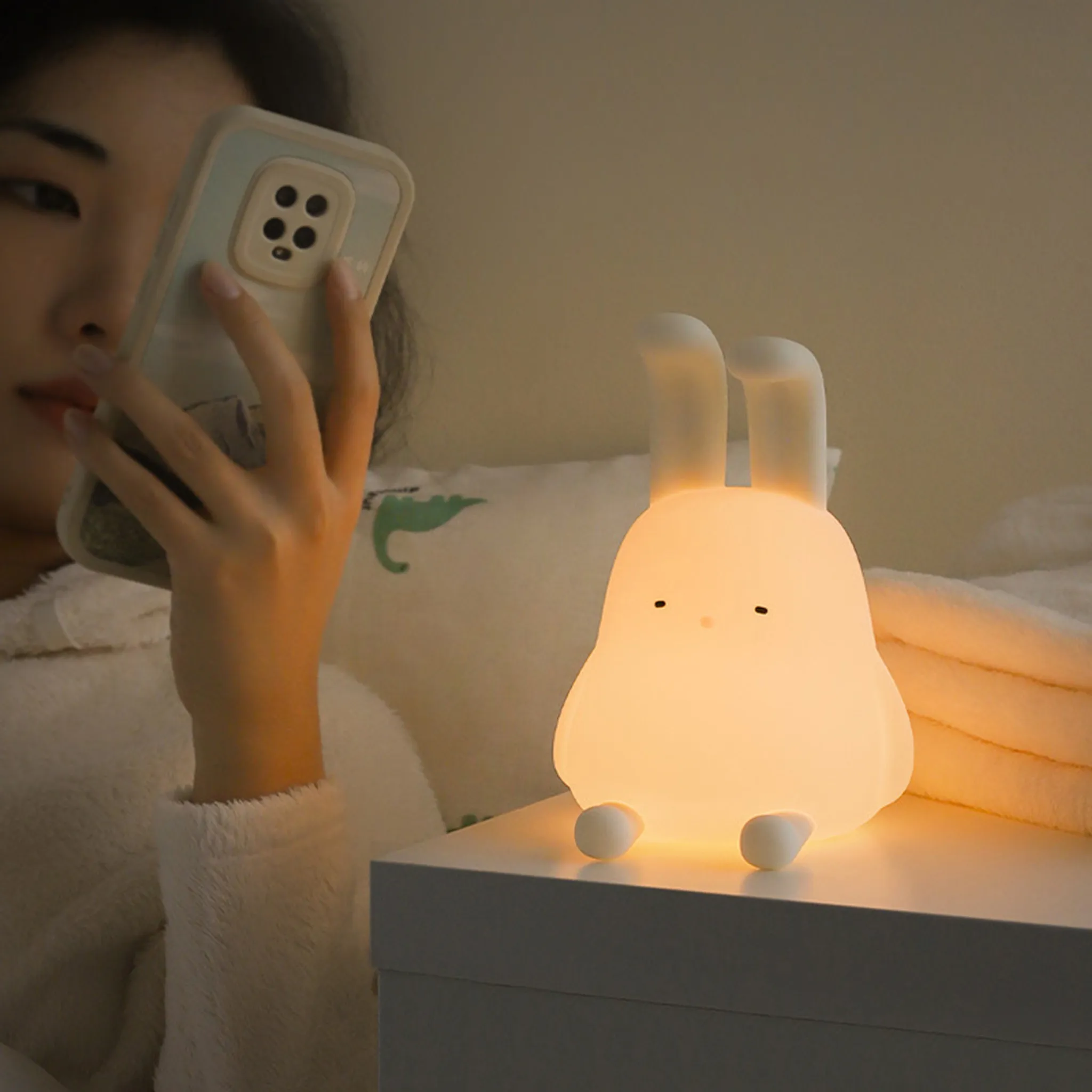 Eine Person benutzt ein Telefon neben einer beleuchteten weißen Hasenlampe auf einem weißen Nachttisch.