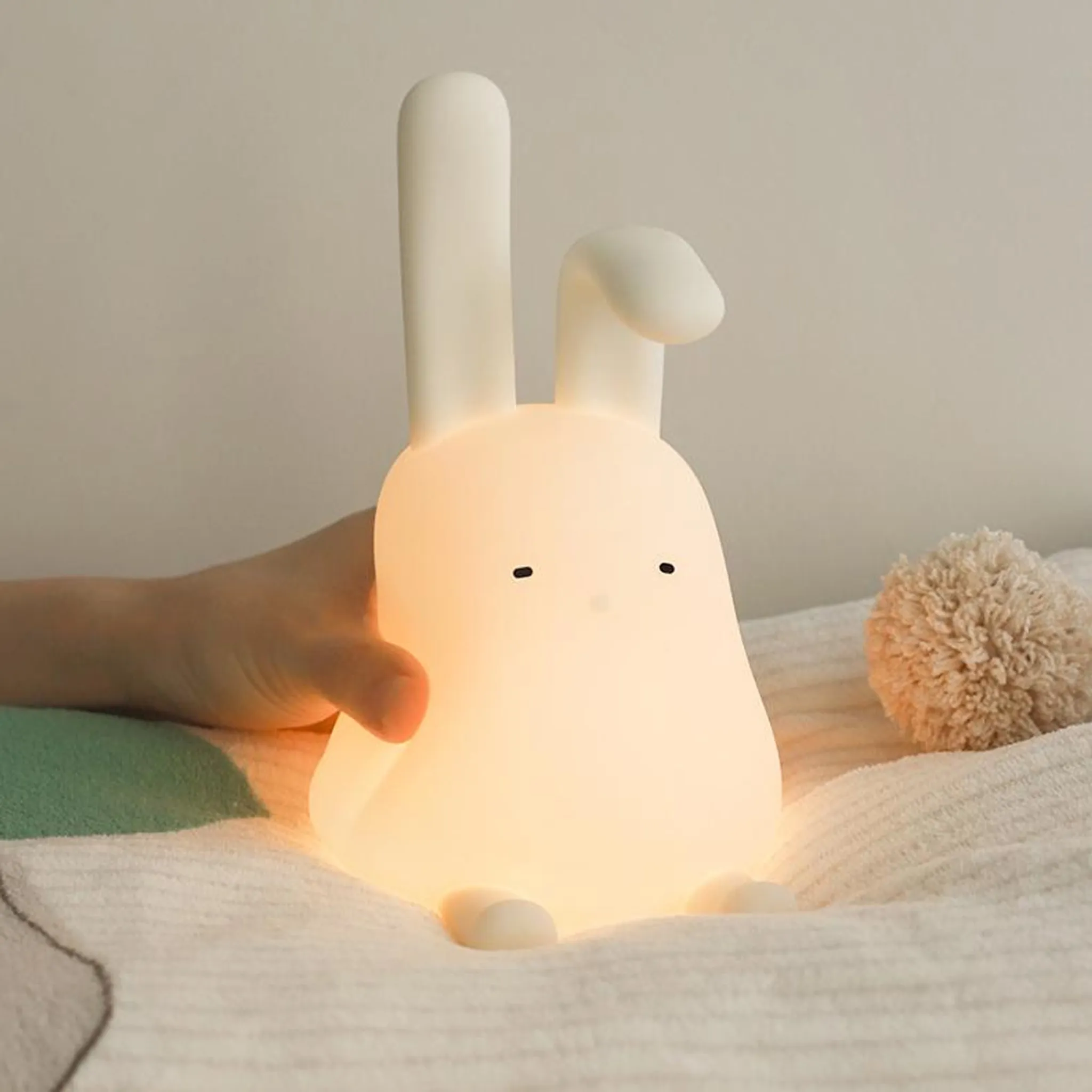 Eine beleuchtete weiße Hasenlampe mit einem Kissen und einer Hand.