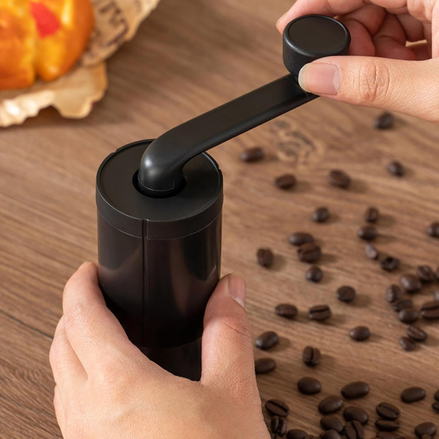 Hände bedienen eine schwarze Kaffeemühle mit Kaffeebohnen auf einem Holztisch.