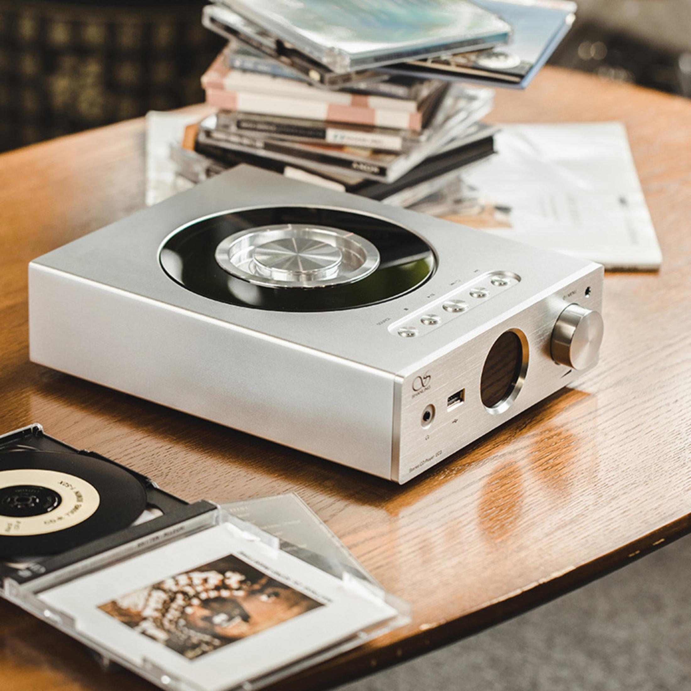 Reproductor de CD plateado con CDs y una caja de CD abierta sobre mesa de madera.