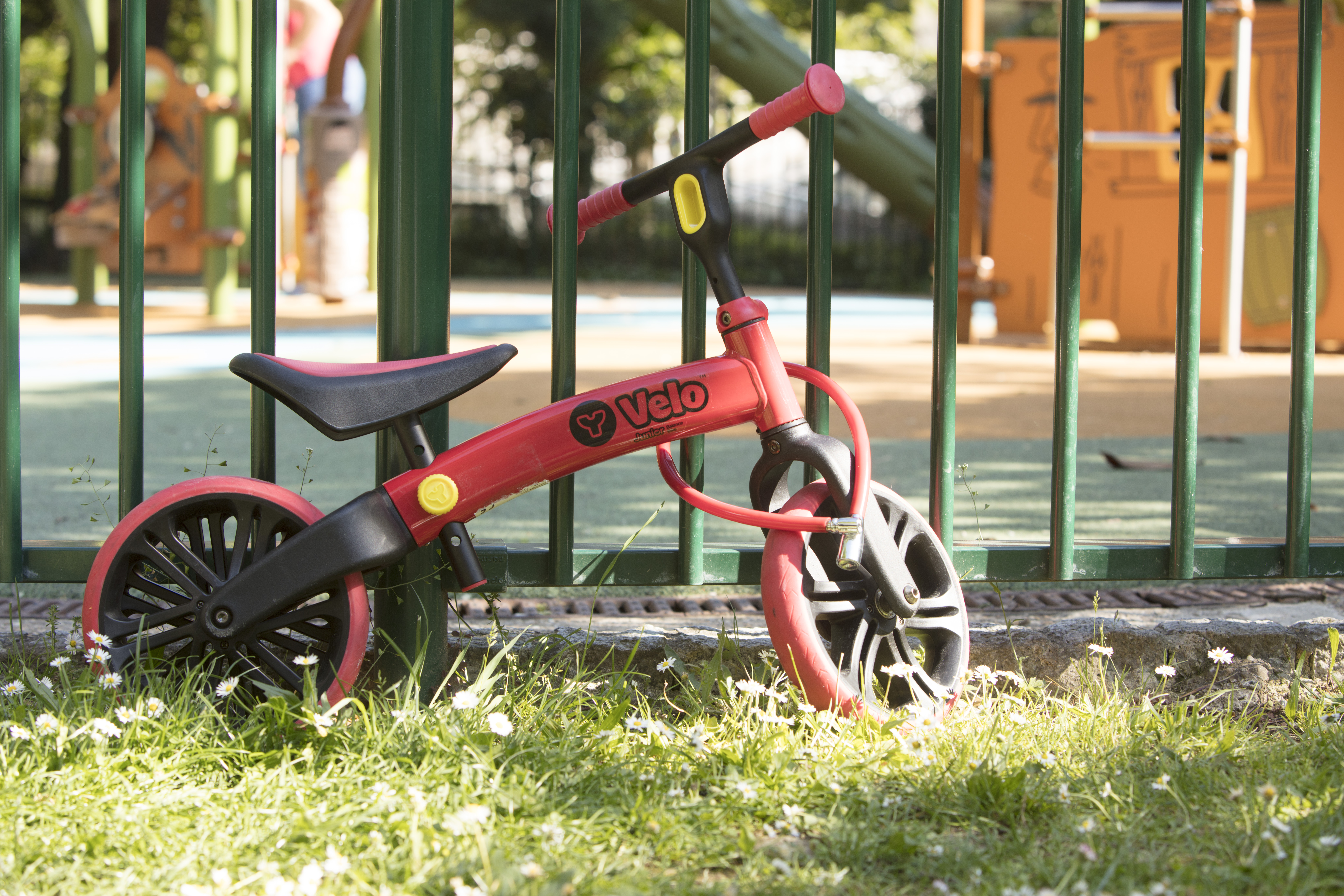 Una bici senza pedali rossa con ruote e manubrio neri è sull'erba davanti a una recinzione.