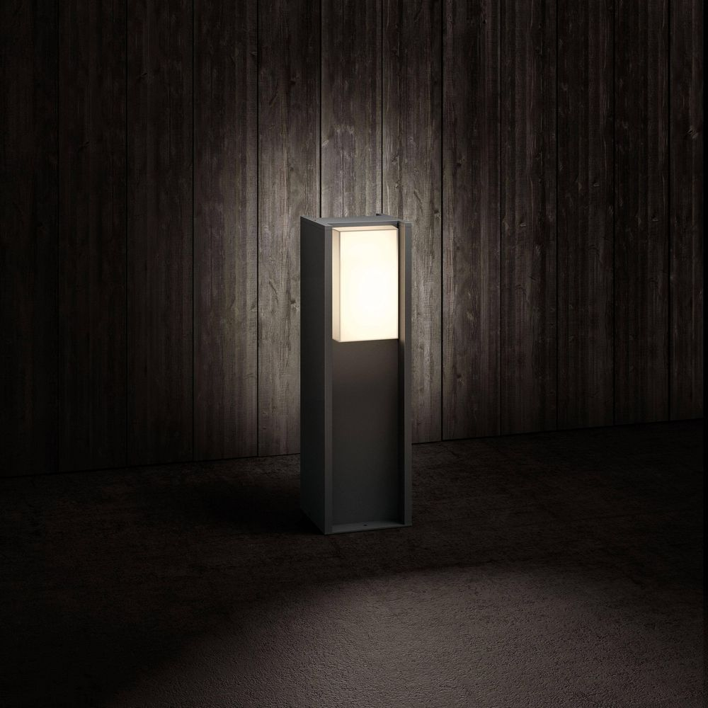 Nowoczesna, ciemna lampa zewnętrzna z podświetlanym prostokątnym kloszem stoi na tle drewnianej ściany.
