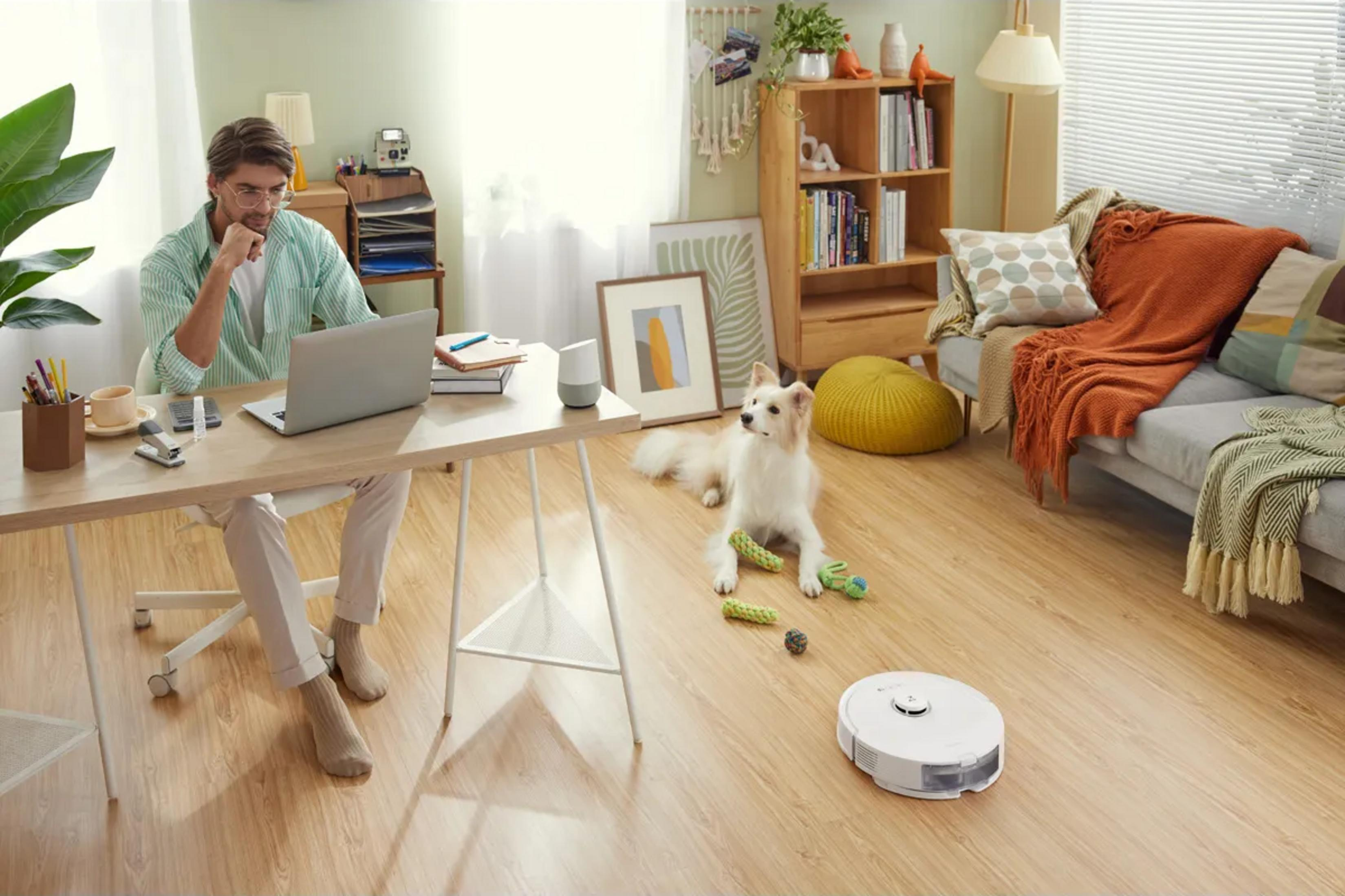 Hombre trabaja en escritorio, perro en suelo, robot aspirador limpia.