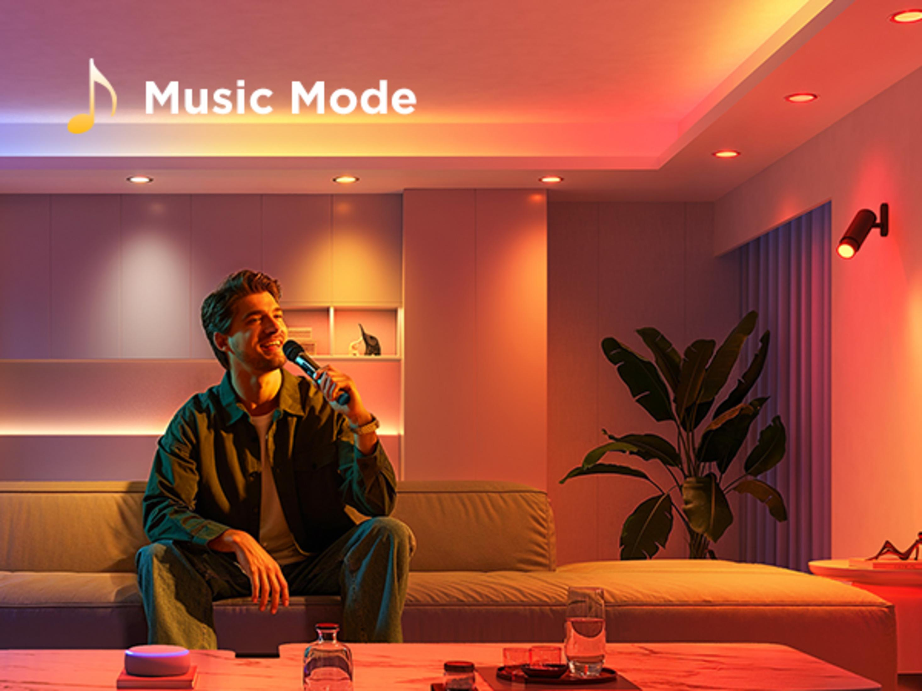 Un uomo seduto su un divano canta in un microfono, illuminato da luci colorate.