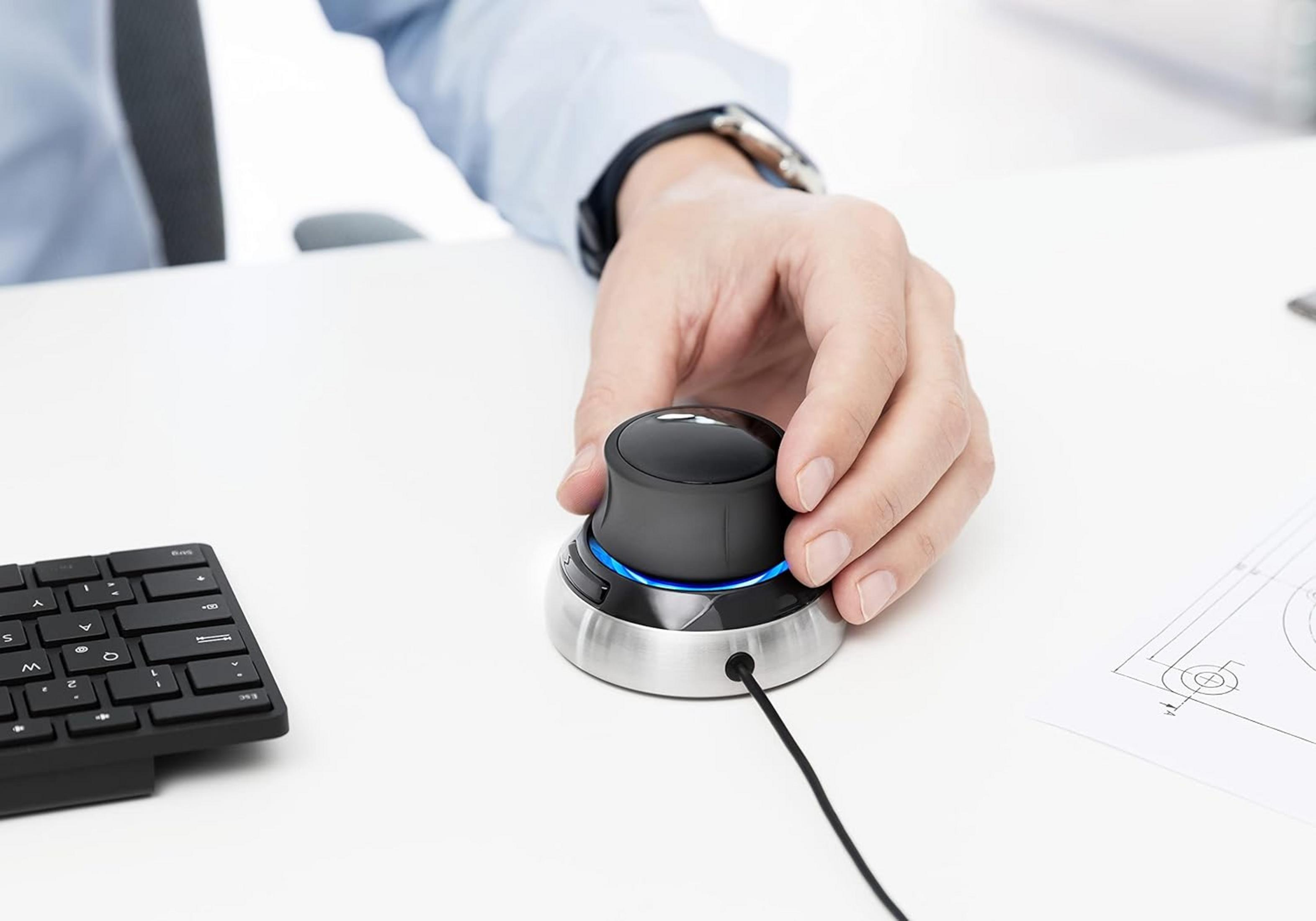 Eine Hand bedient eine 3D-Maus neben einer Tastatur und einem technischen Entwurf.