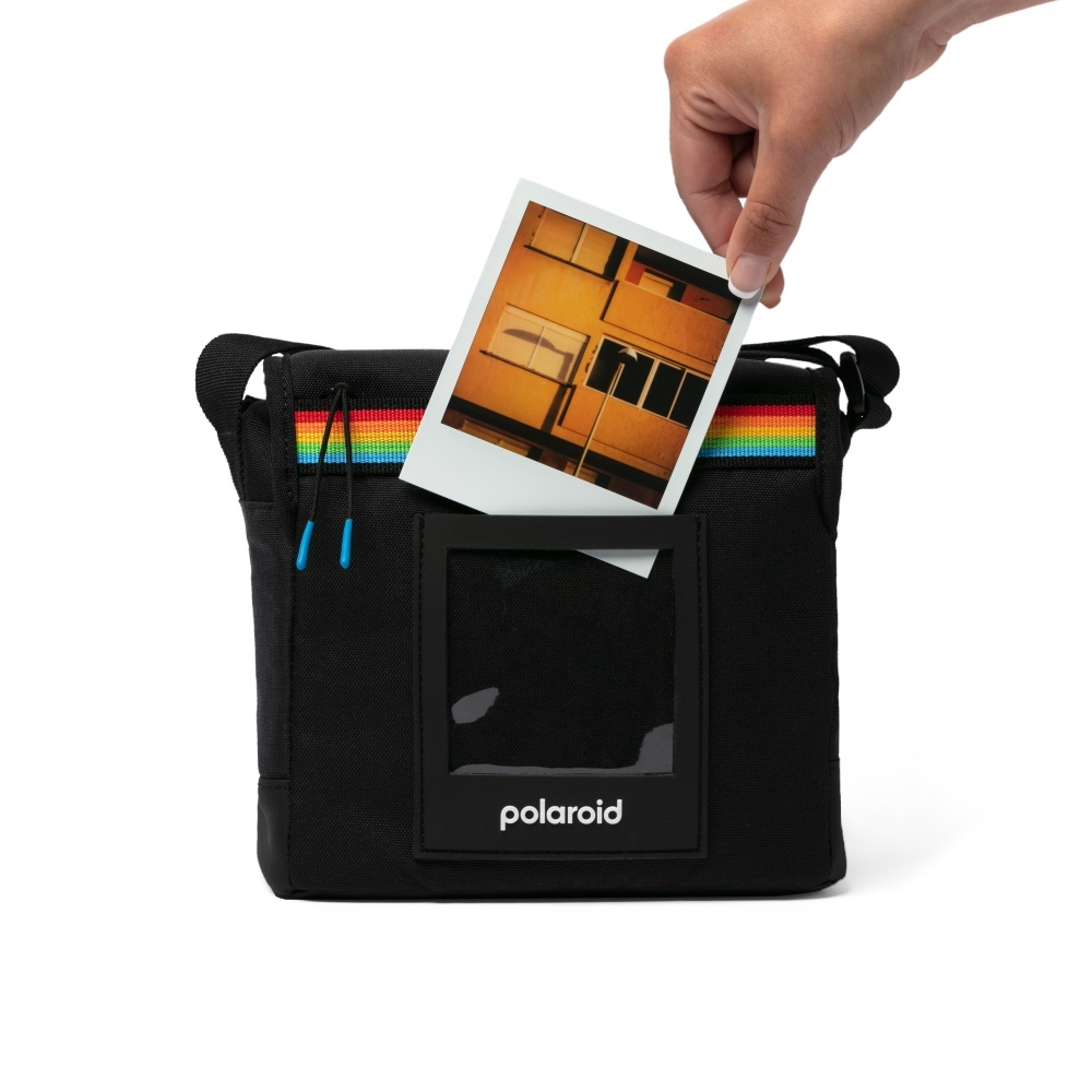 Una mano inserisce una foto Polaroid in una borsa nera a strisce arcobaleno.