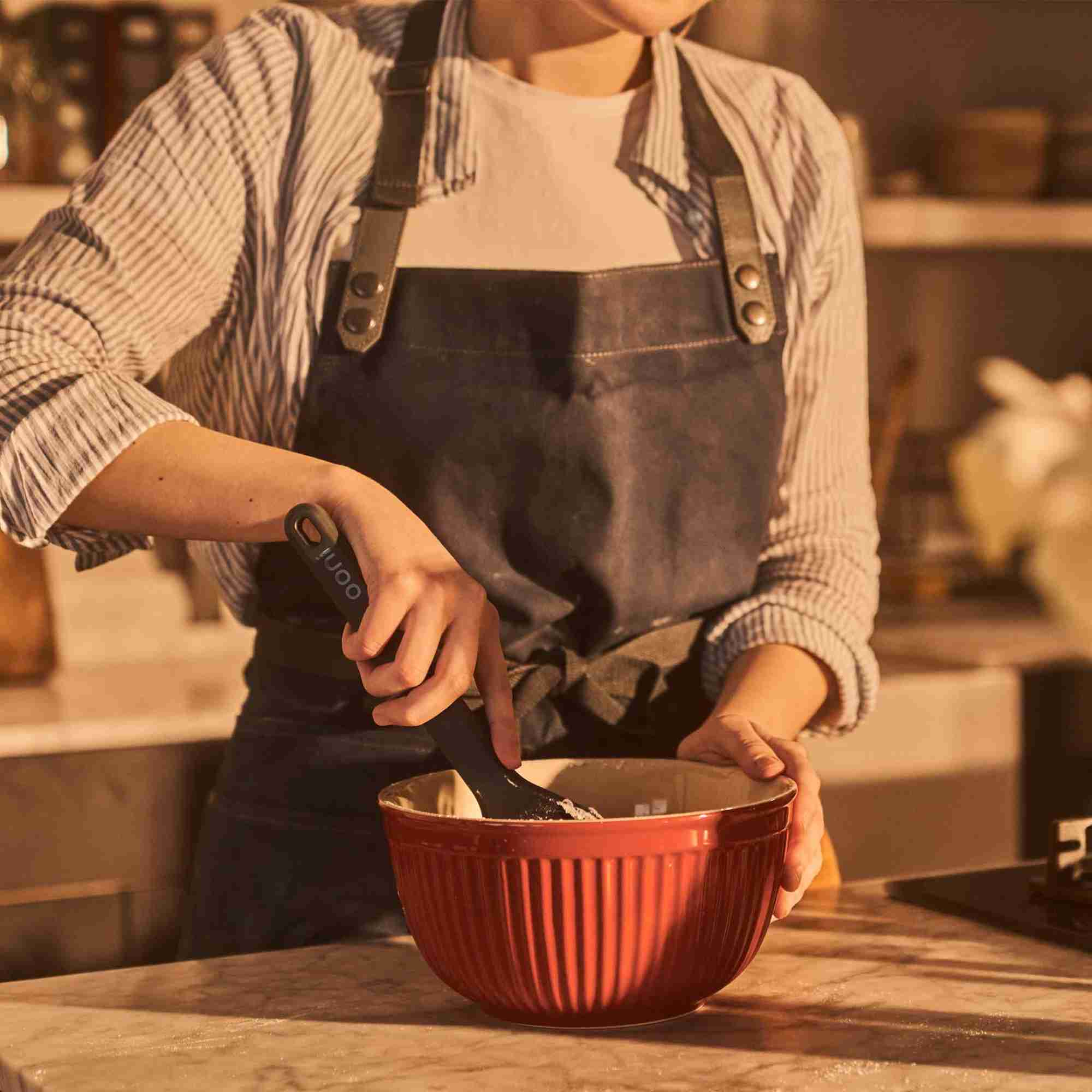 Eine Person in einer Schürze rührt Teig in einer roten Schüssel mit einem schwarzen Utensil.