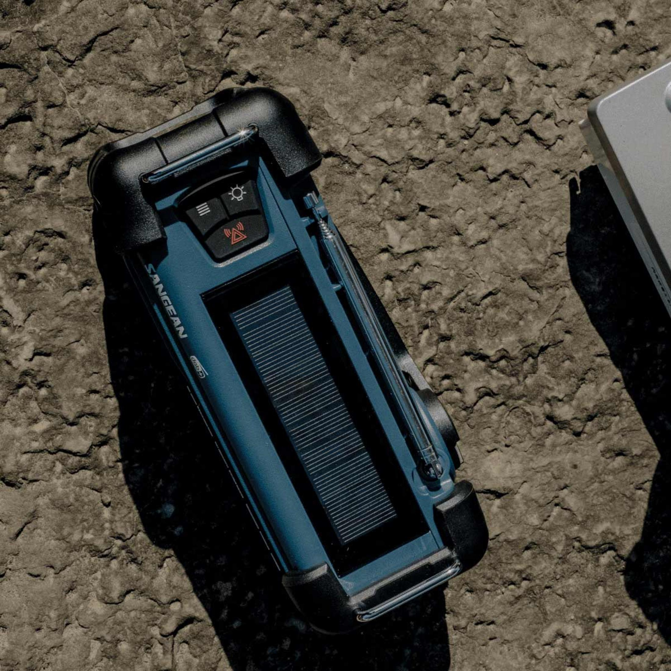 Niebieskie radio Sangean z panelem słonecznym leży na szorstkim, szarym kamieniu.