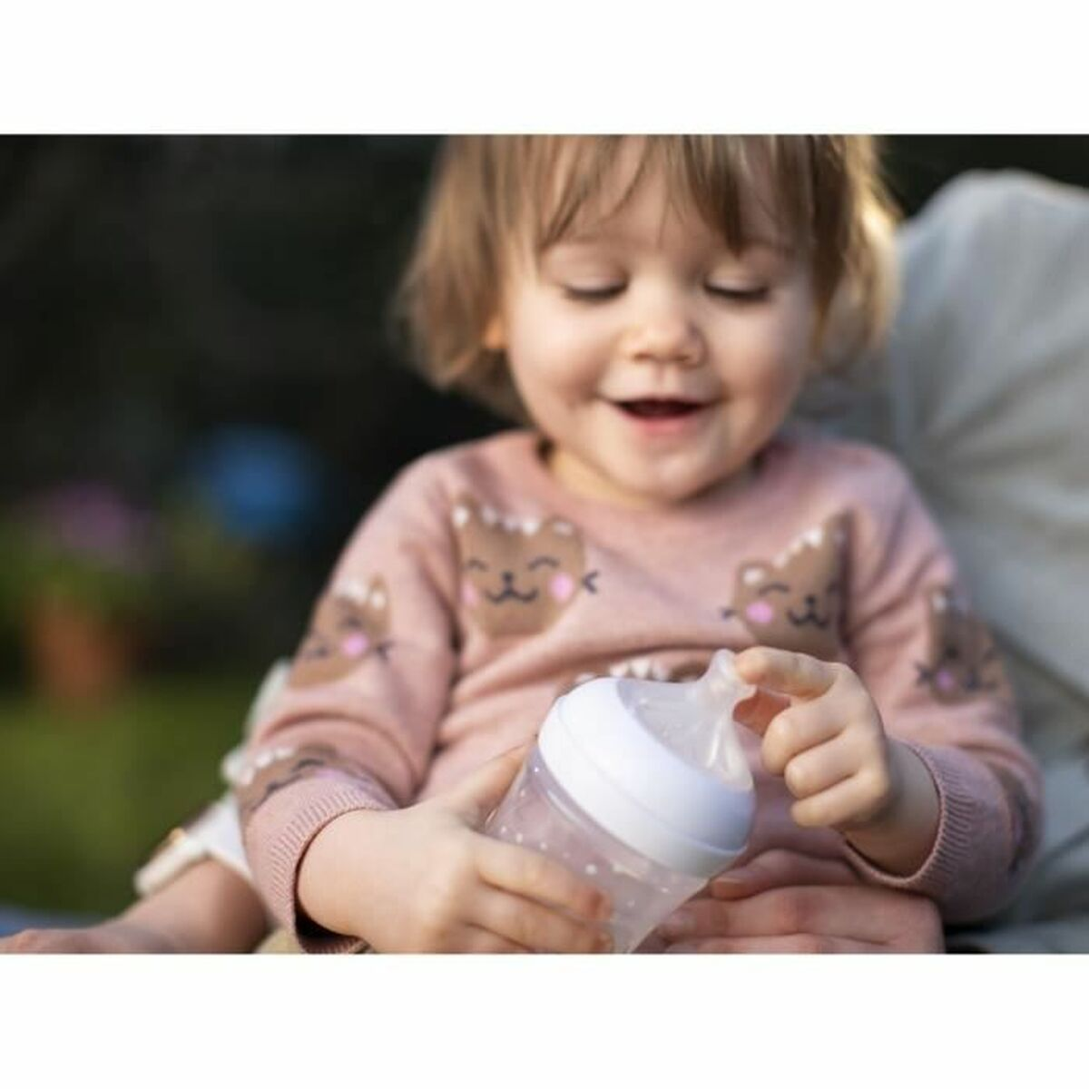 Ein lächelndes Kleinkind in einem rosafarbenen Pullover mit Teddybären hält eine Babyflasche.