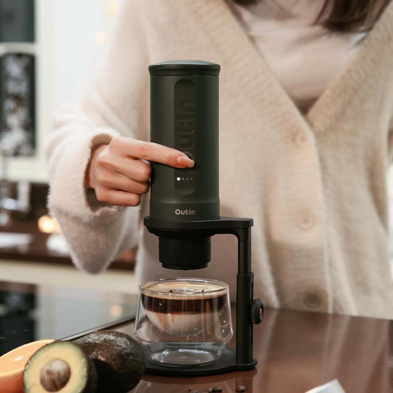 Una persona opera una cafetera, goteando café en un vaso. Hay fruta cerca.