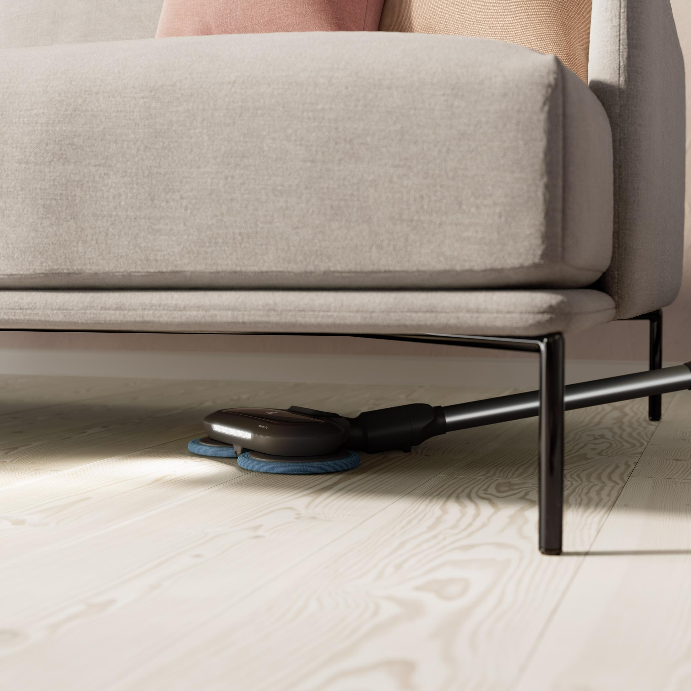 Un robot aspirapolvere con spazzole blu pulisce sotto un divano grigio su un pavimento in legno chiaro.