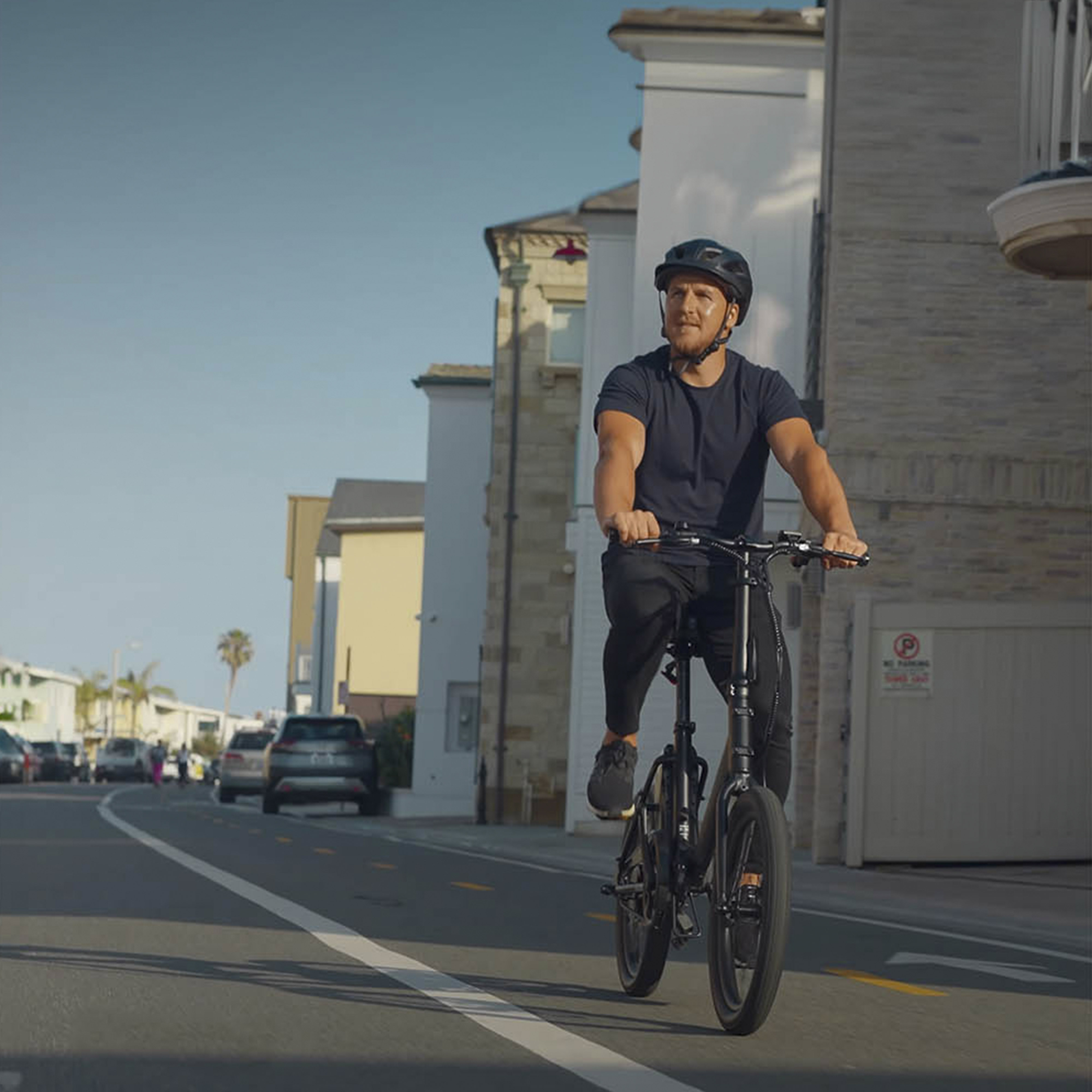 Un hombre monta en bicicleta por una calle, con casas y coches de fondo.