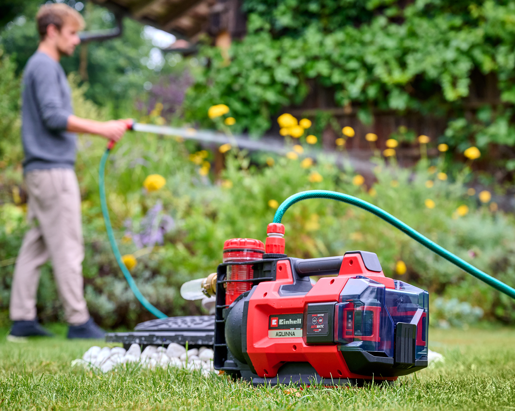 Ein Mann bewässert einen Garten mit einer roten Pumpe und einem grünen Schlauch im Vordergrund.