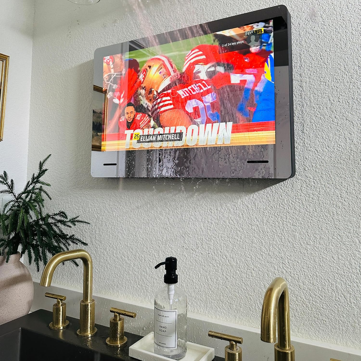 Ein an der Wand montierter Fernseher zeigt ein Footballspiel. Darunter ist ein Waschbecken mit Wasserhähnen.