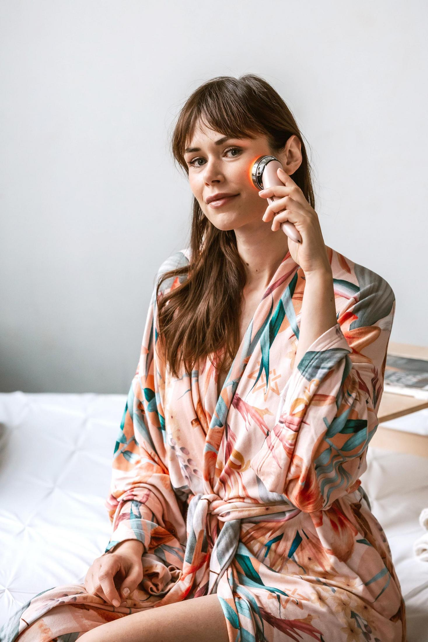 Kobieta używa urządzenia do twarzy, nosi szlafrok w kwiaty, siedzi wewnątrz.