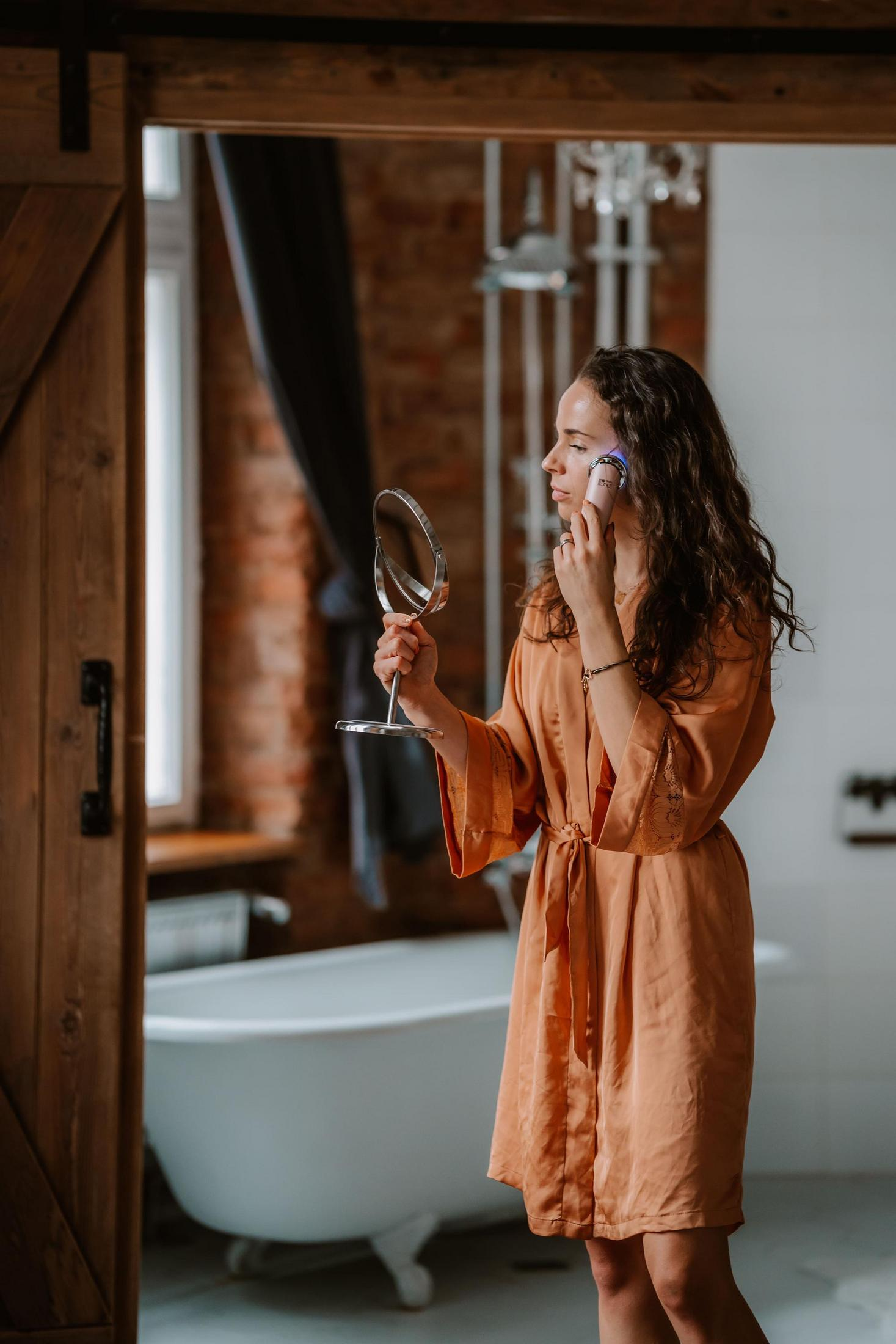 Kobieta w pomarańczowym szlafroku trzyma lustro, używa urządzenia do twarzy. Wanna w tle.