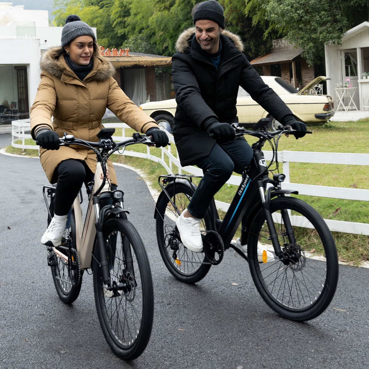 Mężczyzna i kobieta jadą rowerami po utwardzonej drodze z białym ogrodzeniem i drzewami.