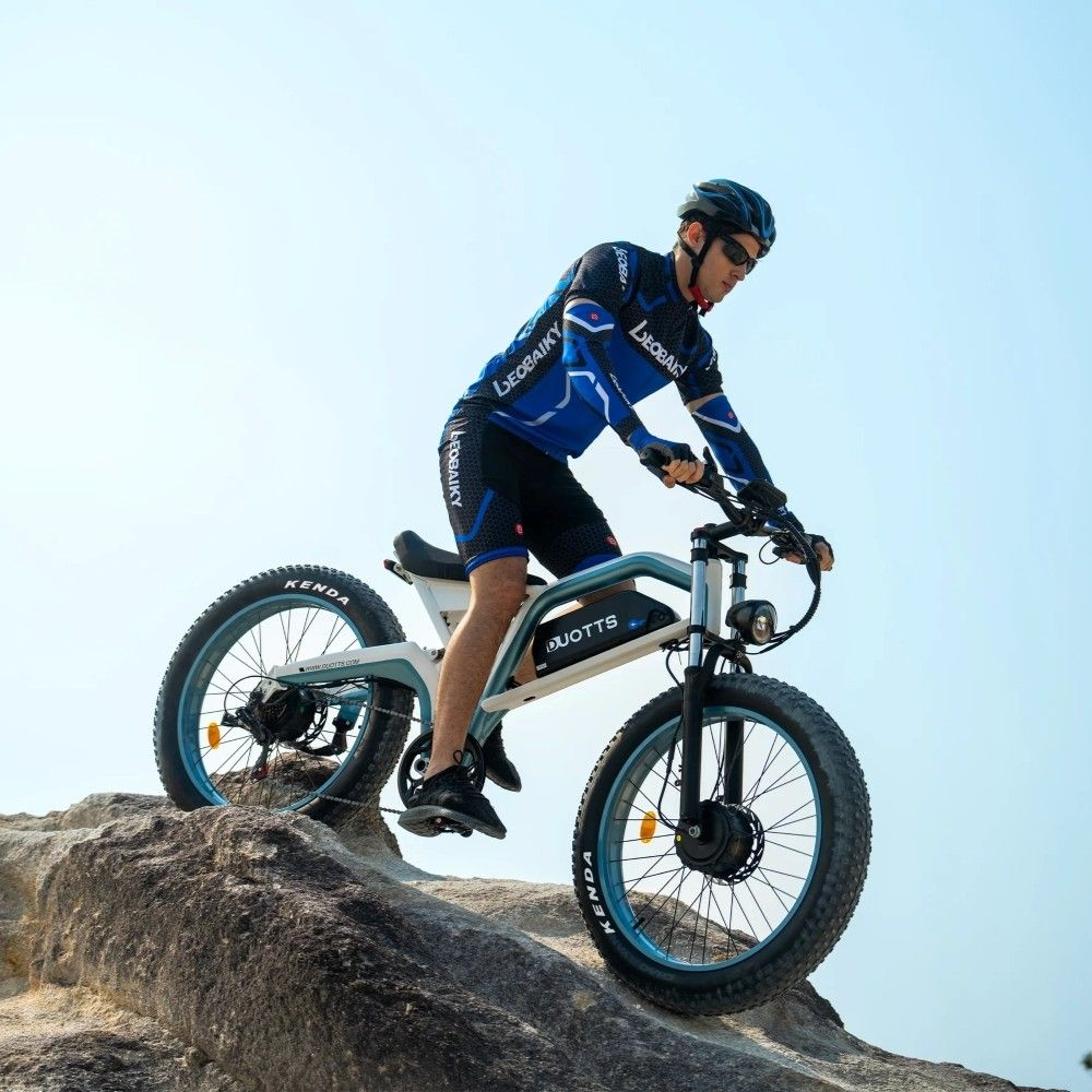 Un uomo in tuta da ciclismo blu guida una e-bike con copertoni larghi su terreno roccioso.