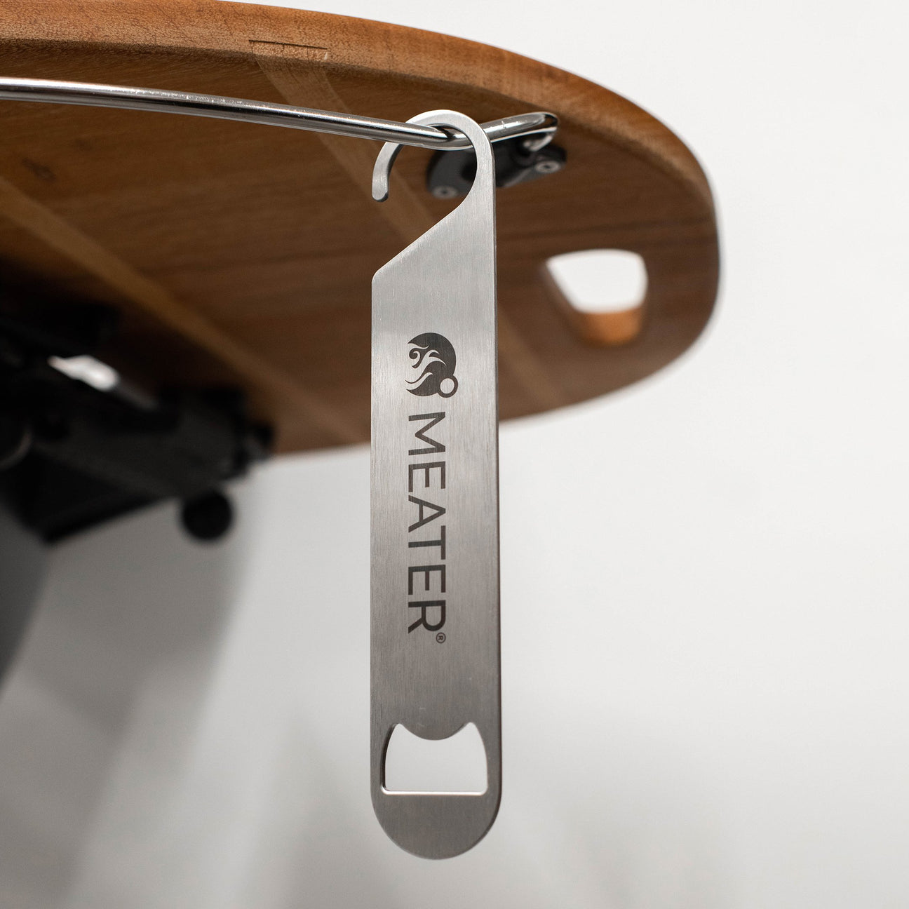 Ein silberner Flaschenöffner mit dem Logo "MEATER" hängt an einem Metallstab unter einem Holzbrett.