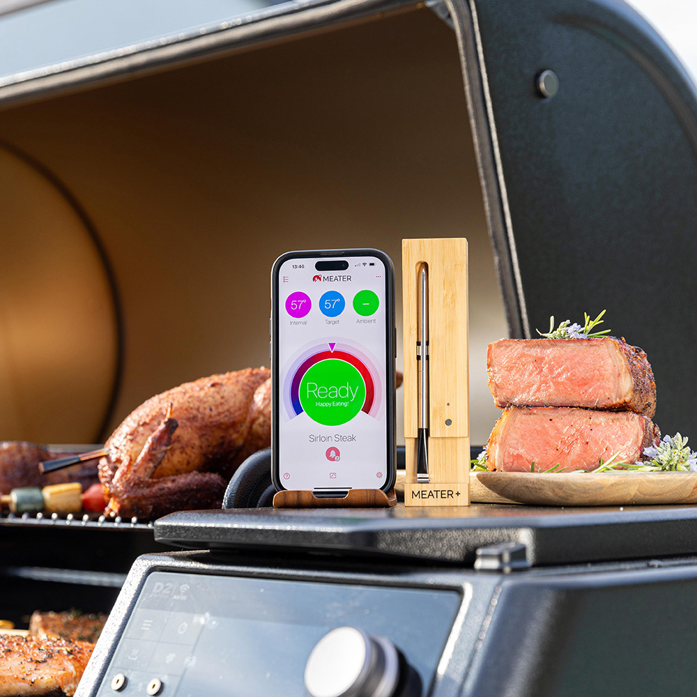 Ein Smartphone zeigt eine Grill-App, daneben ein Fleischthermometer und gegrilltes Fleisch.