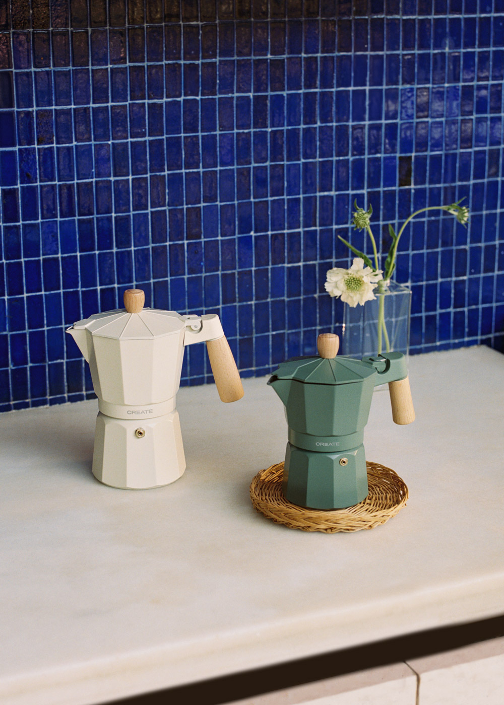 Dos cafeteras moka, una blanca y otra verde, sobre un mostrador de mármol frente a azulejos azules.