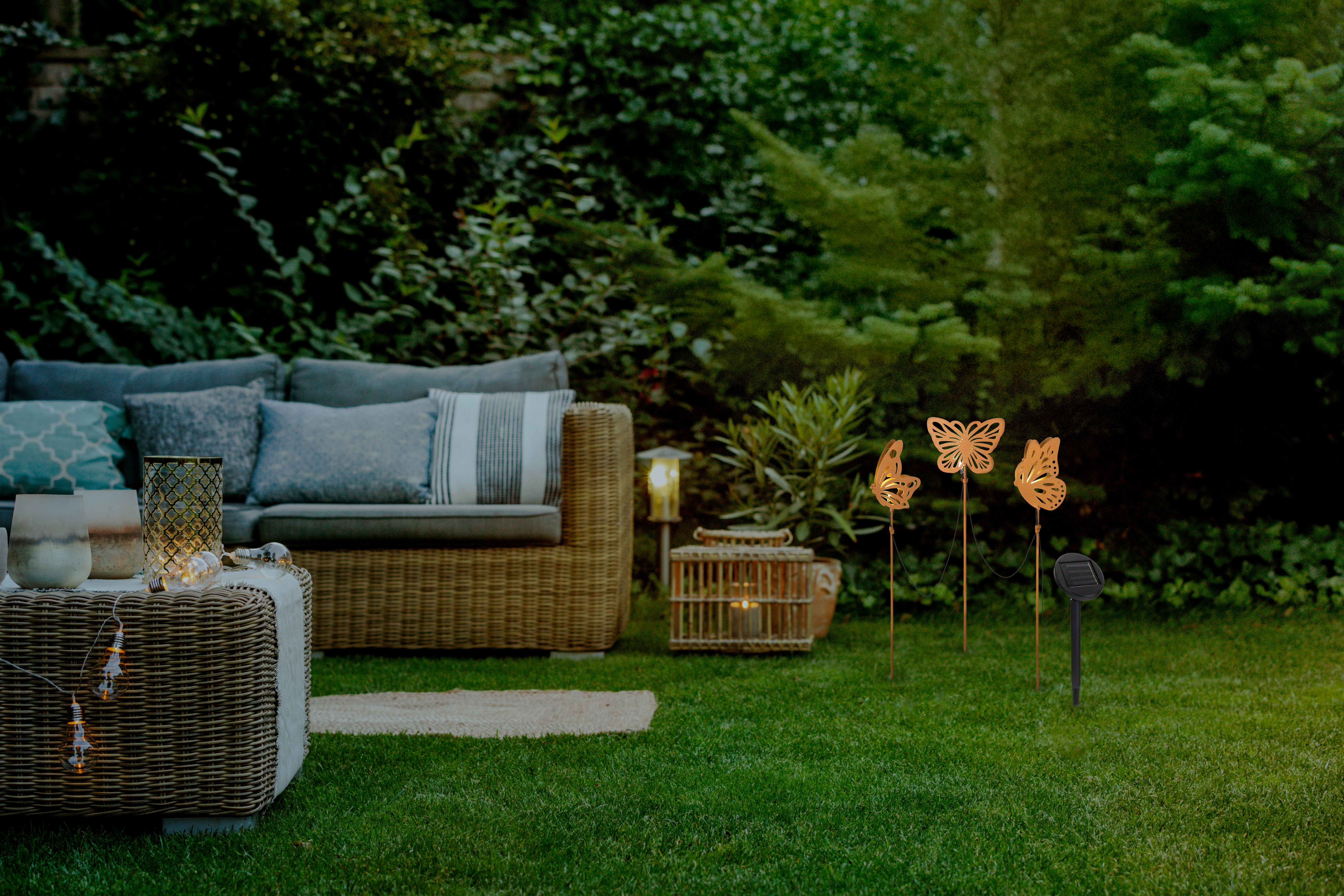 Gartenmöbel mit Kissen, Lichterkette und Schmetterlingslampen bei Nacht.
