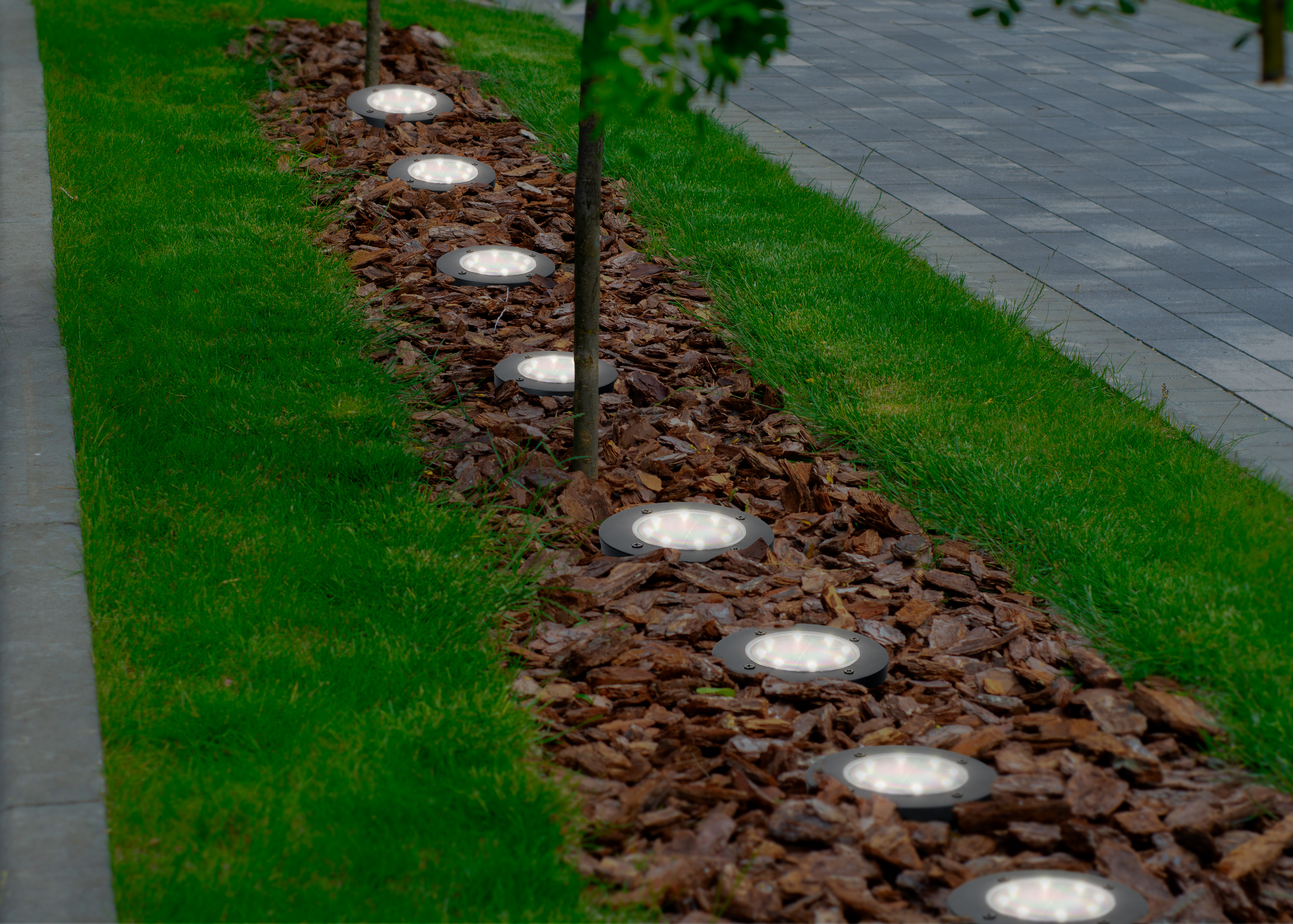 Reihe von Bodeneinbaustrahlern beleuchtet einen Weg aus Rindenmulch neben Gras.