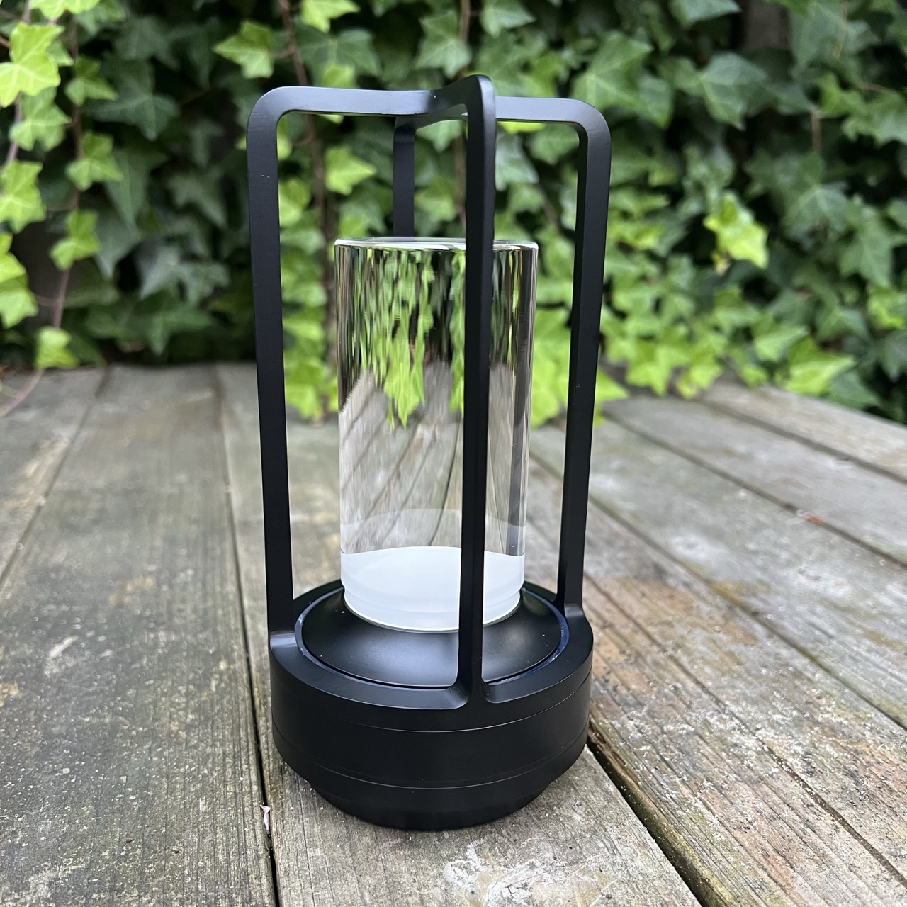 Schwarze Laterne mit Glaszylinder auf Holzoberfläche vor Efeubewuchs.