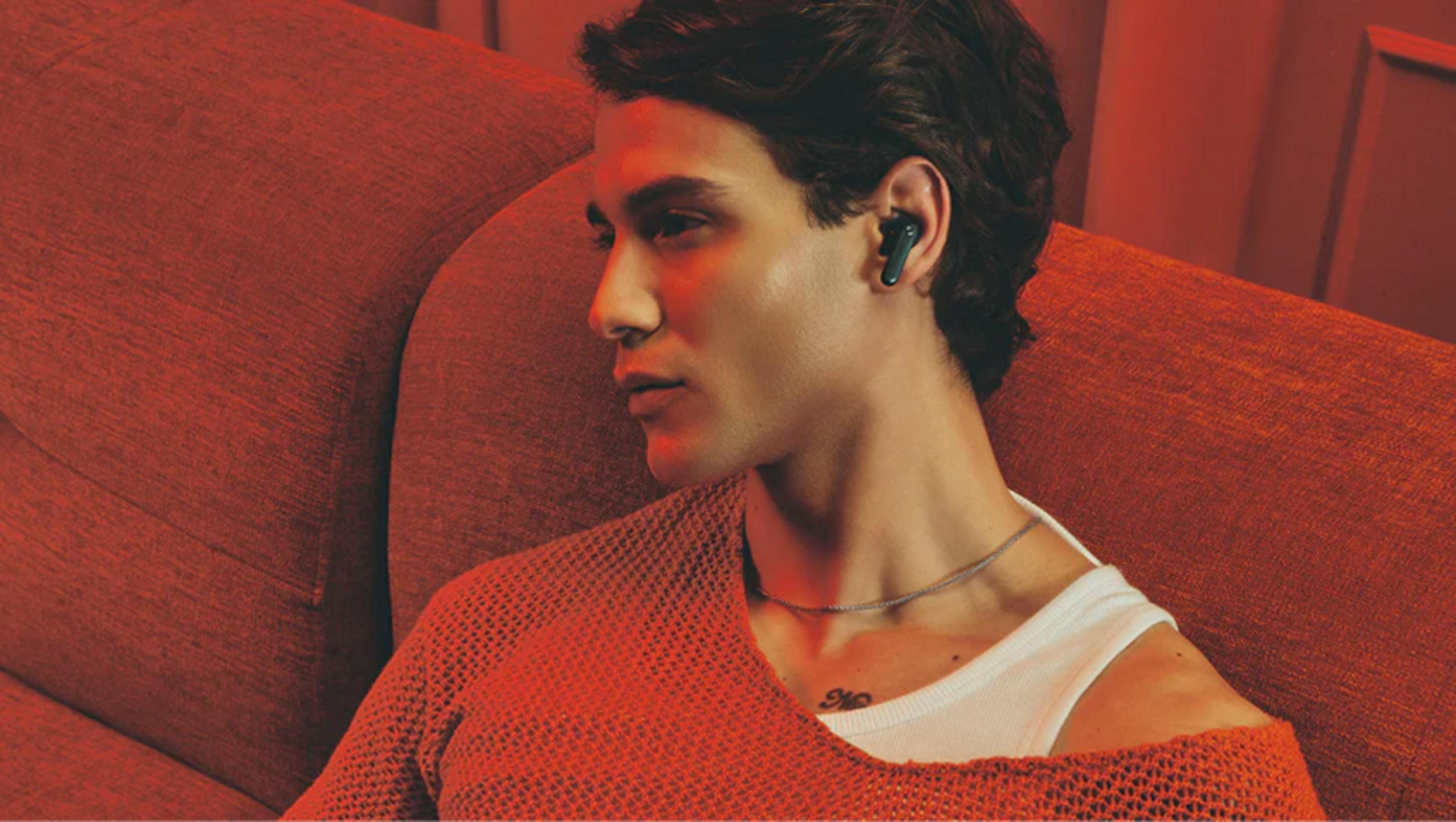 Mężczyzna z słuchawką siedzi na czerwonej kanapie, w pomarańczowym swetrze.