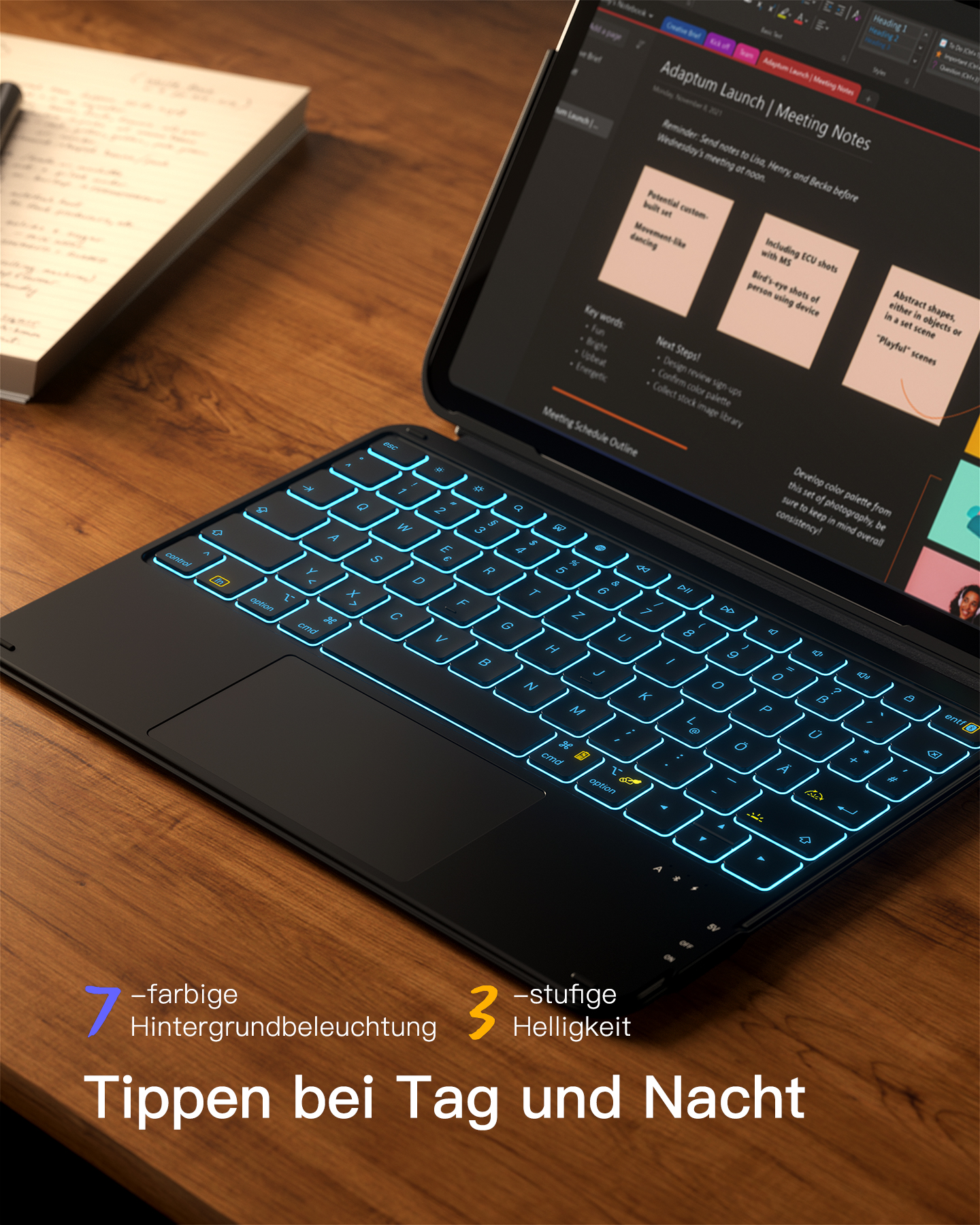 Ein Laptop mit blau beleuchteter Tastatur auf einem Holztisch. Notizbuch daneben.