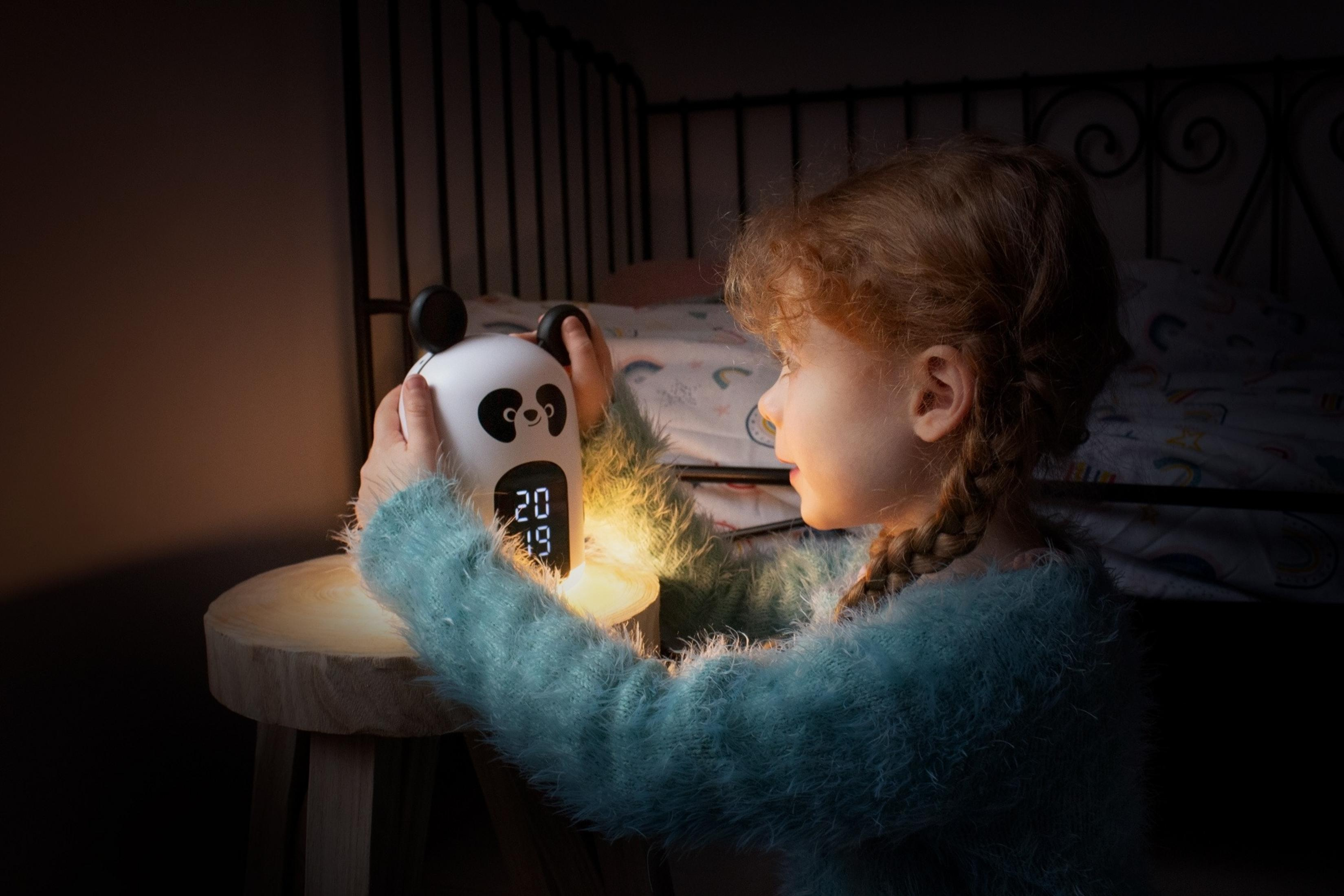 Dziewczynka z rudymi włosami trzyma budzik panda w ciemnym pokoju.
