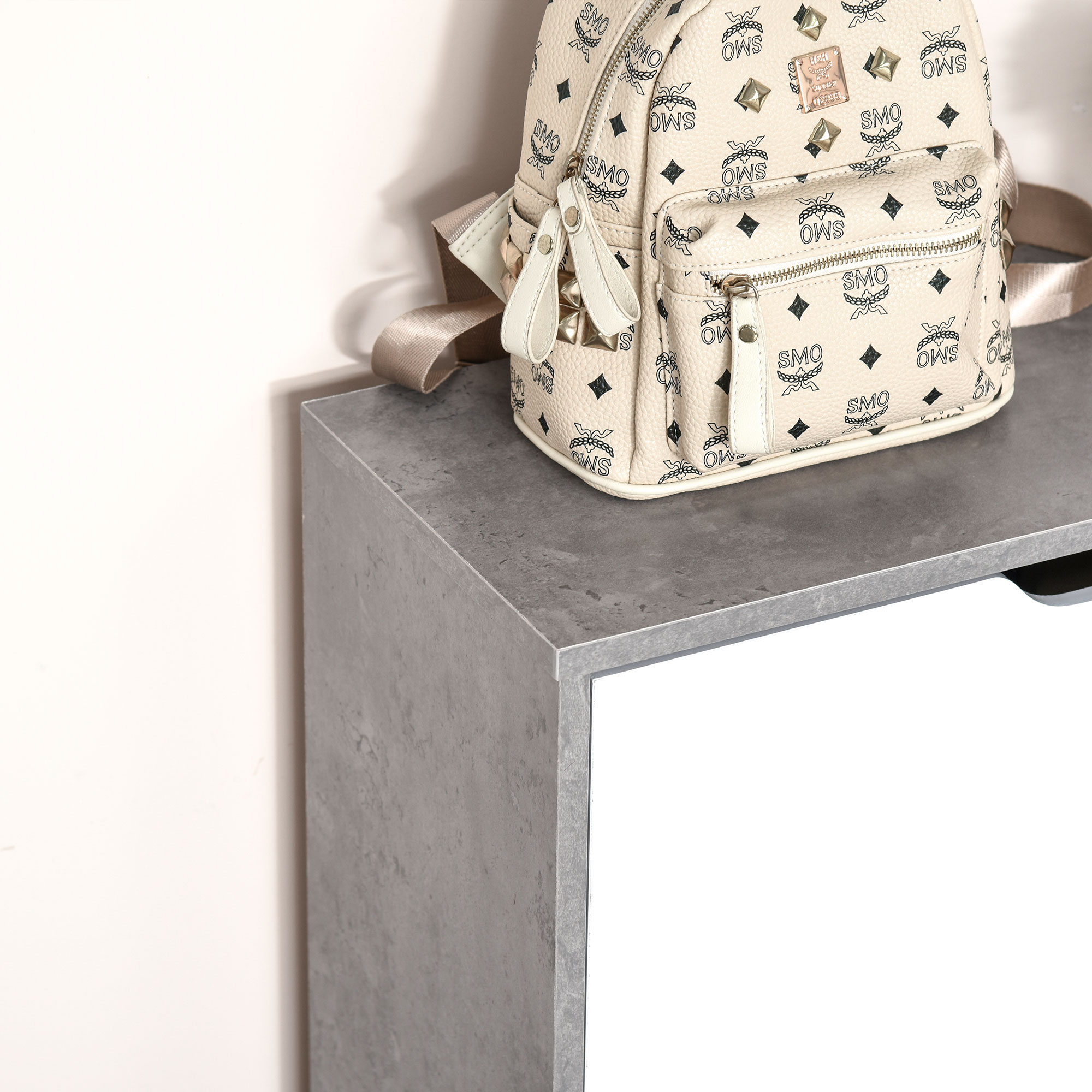 Una mochila beige con estampado de logo negro y tachuelas está sobre una mesa gris.