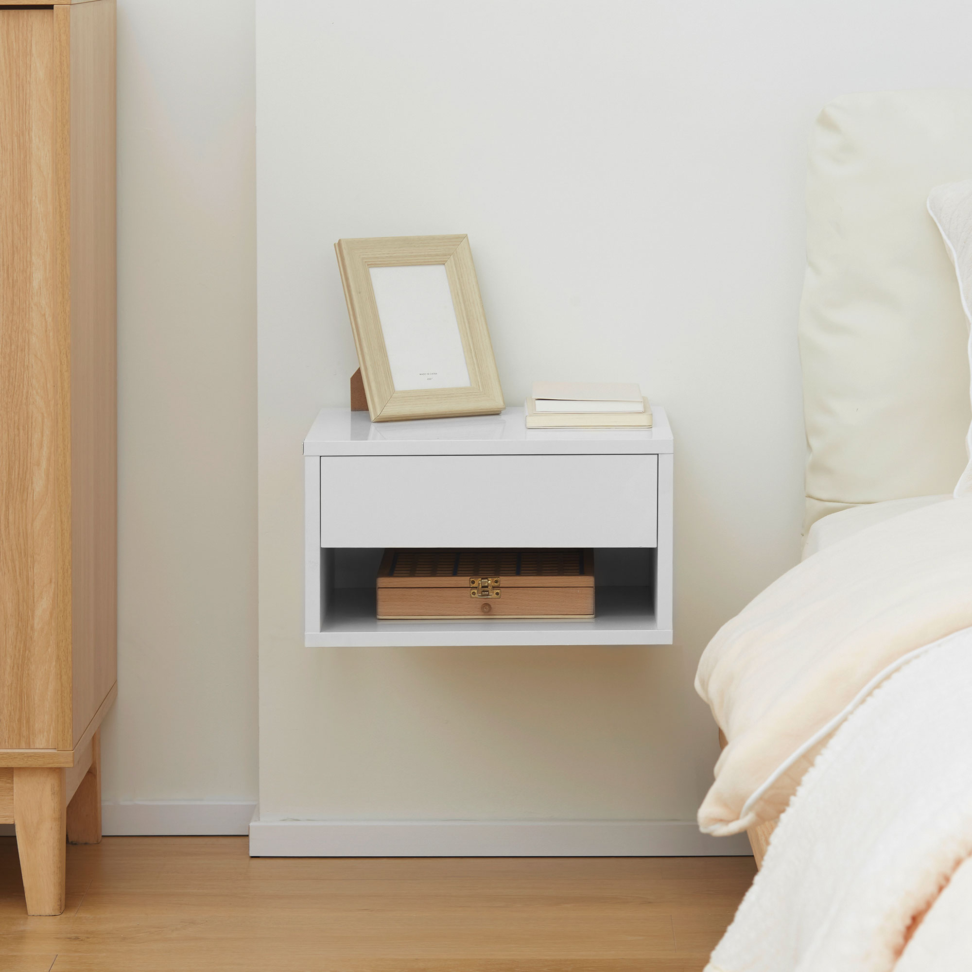 Una mesita de noche flotante blanca con un marco de fotos de madera, dos libros y una caja de madera en una pared clara junto a una cama.