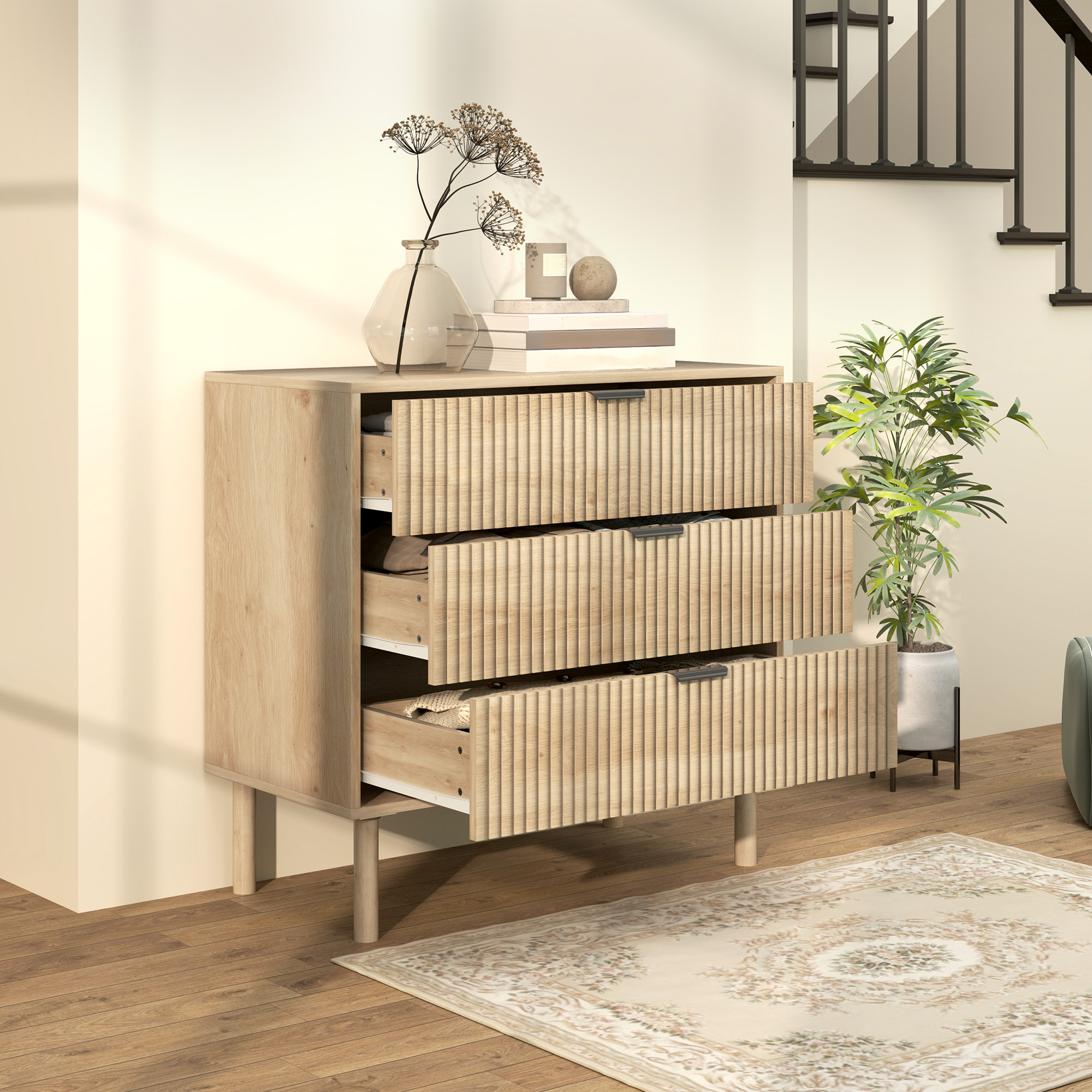 Cómoda de madera clara con tres cajones sobre suelo de madera junto a escaleras.