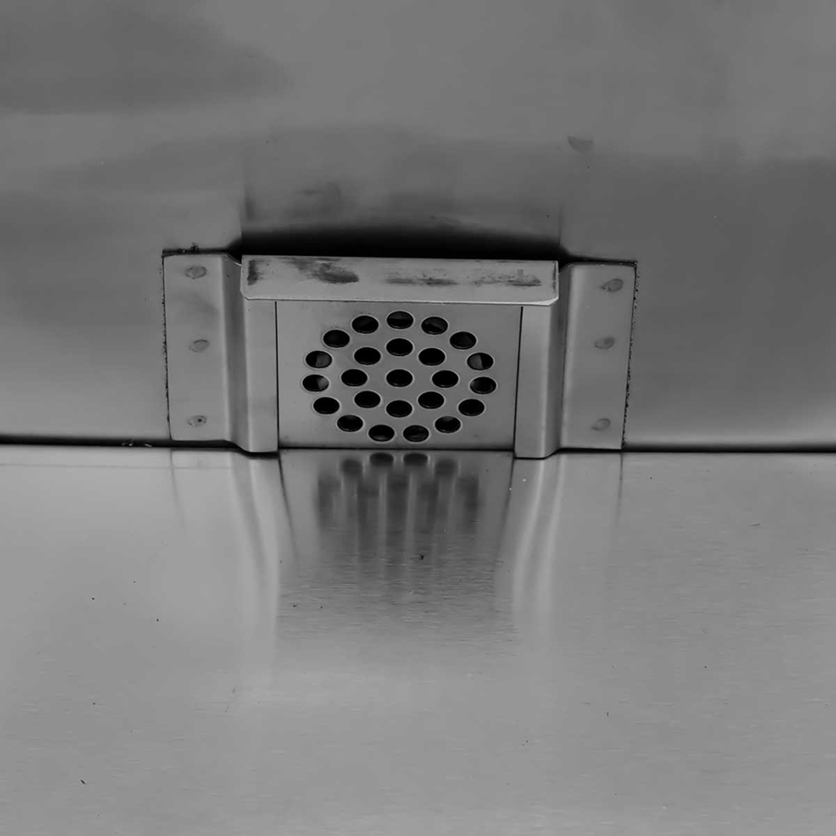 A rectangular vent grille with many round holes, mounted on a metal surface.