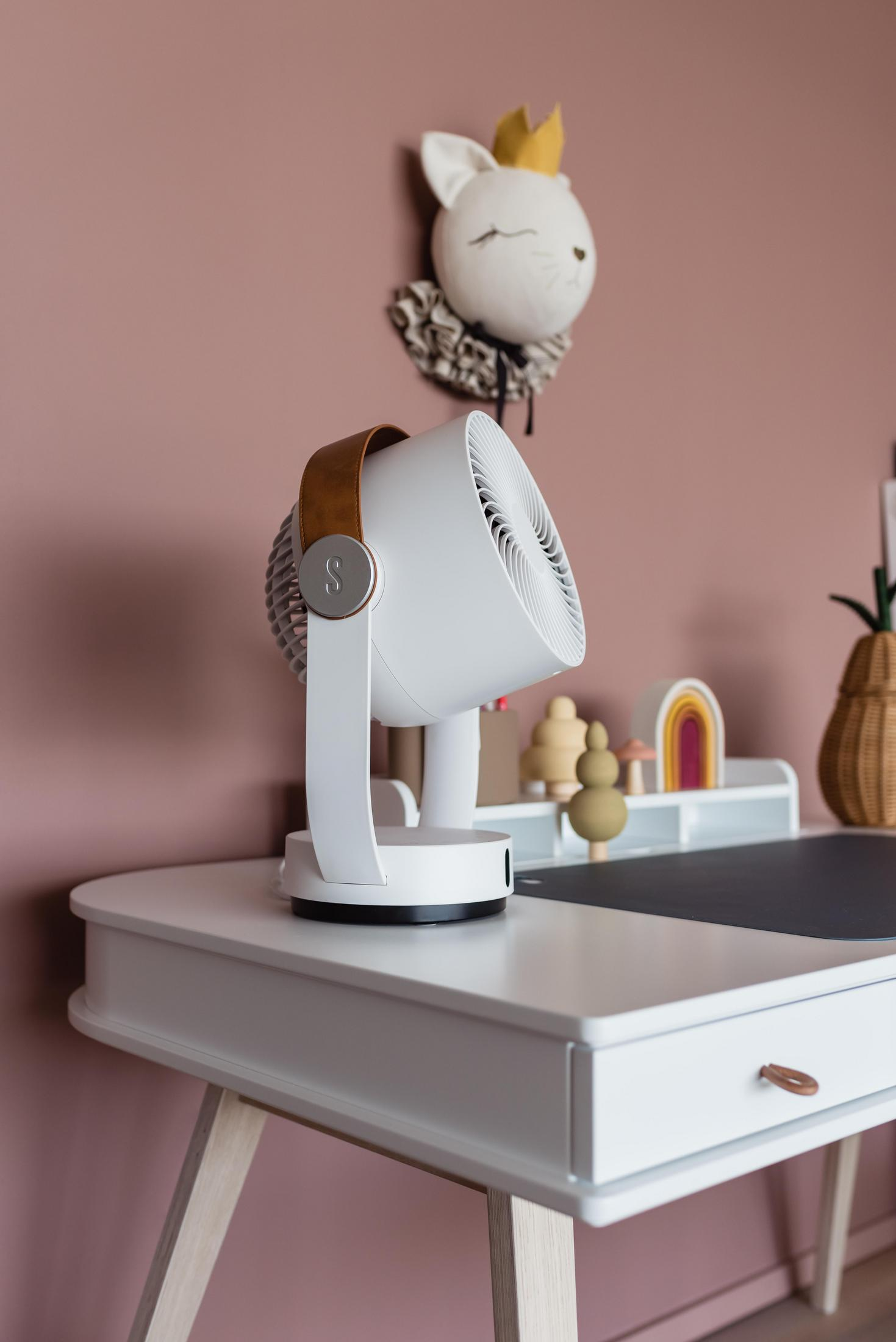 Ein weißer Ventilator steht auf einem Schreibtisch vor einer rosa Wand mit Deko.