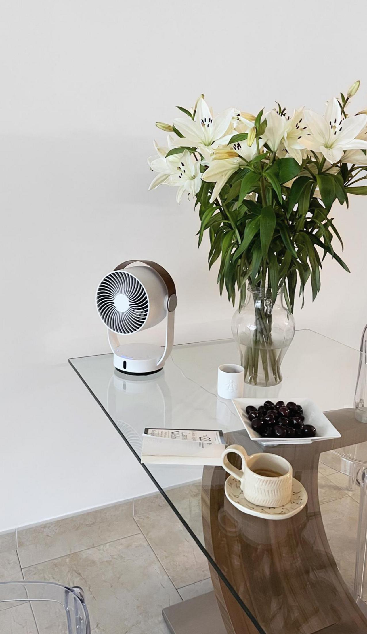 Ein weißer Ventilator, weiße Lilien, Kaffeetasse und Kirschen auf einem Glastisch.