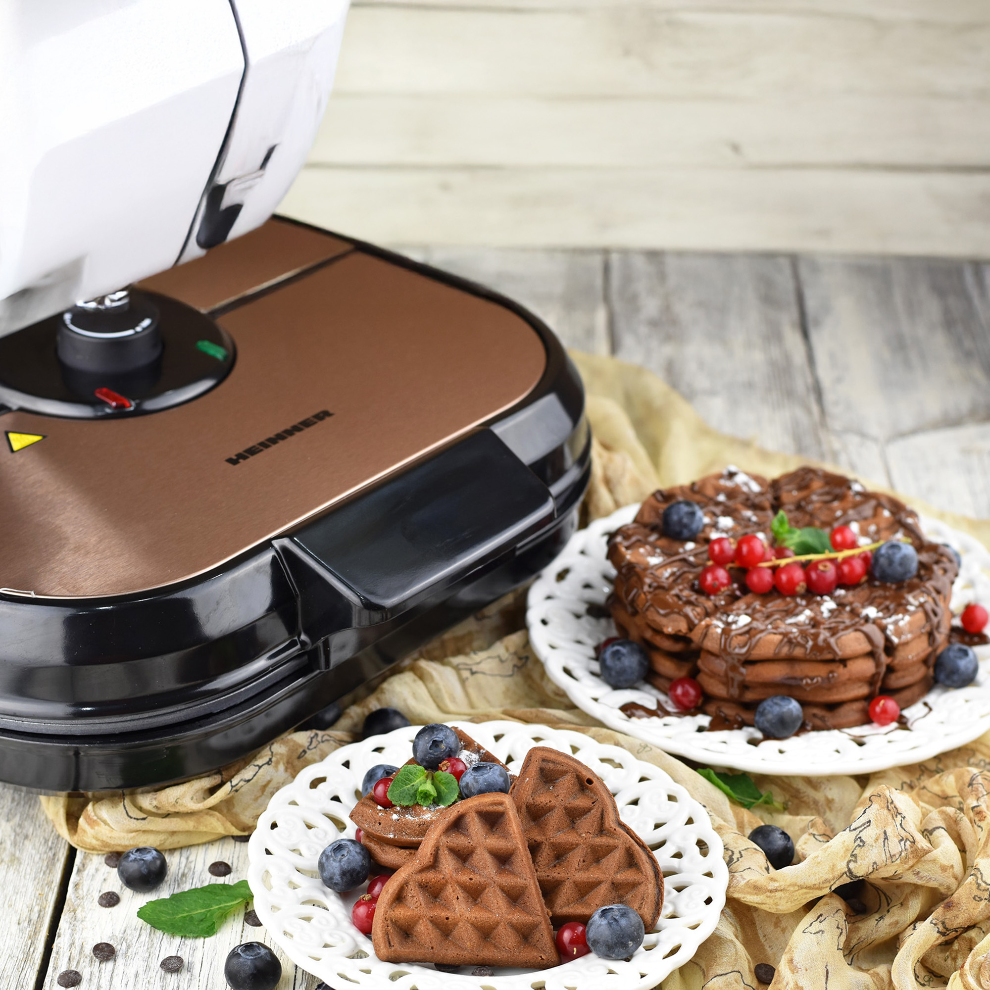 A waffle maker sits beside heart-shaped chocolate waffles with berries and chocolate drizzle.