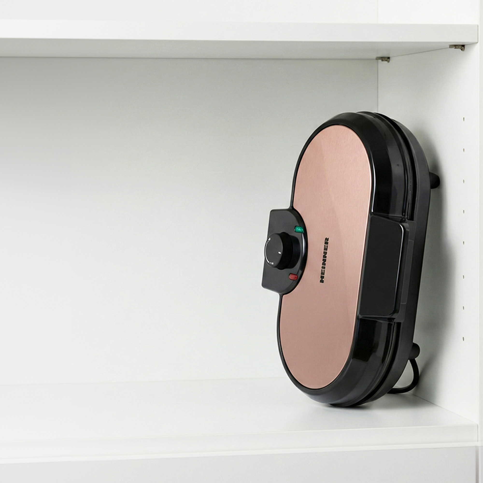 A rose gold waffle maker with a black casing rests inside a white shelf.