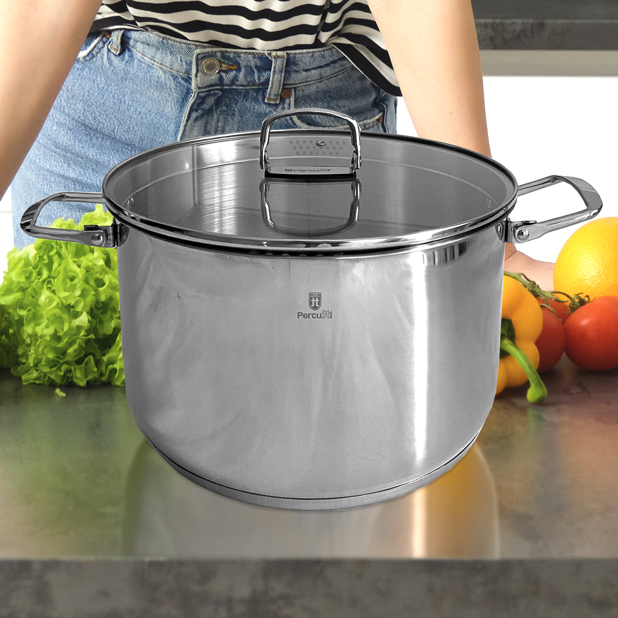 Mujer con jeans y top a rayas sostiene una olla de acero inoxidable con tapa. Al lado hay lechuga, limón, tomates y pimiento.
