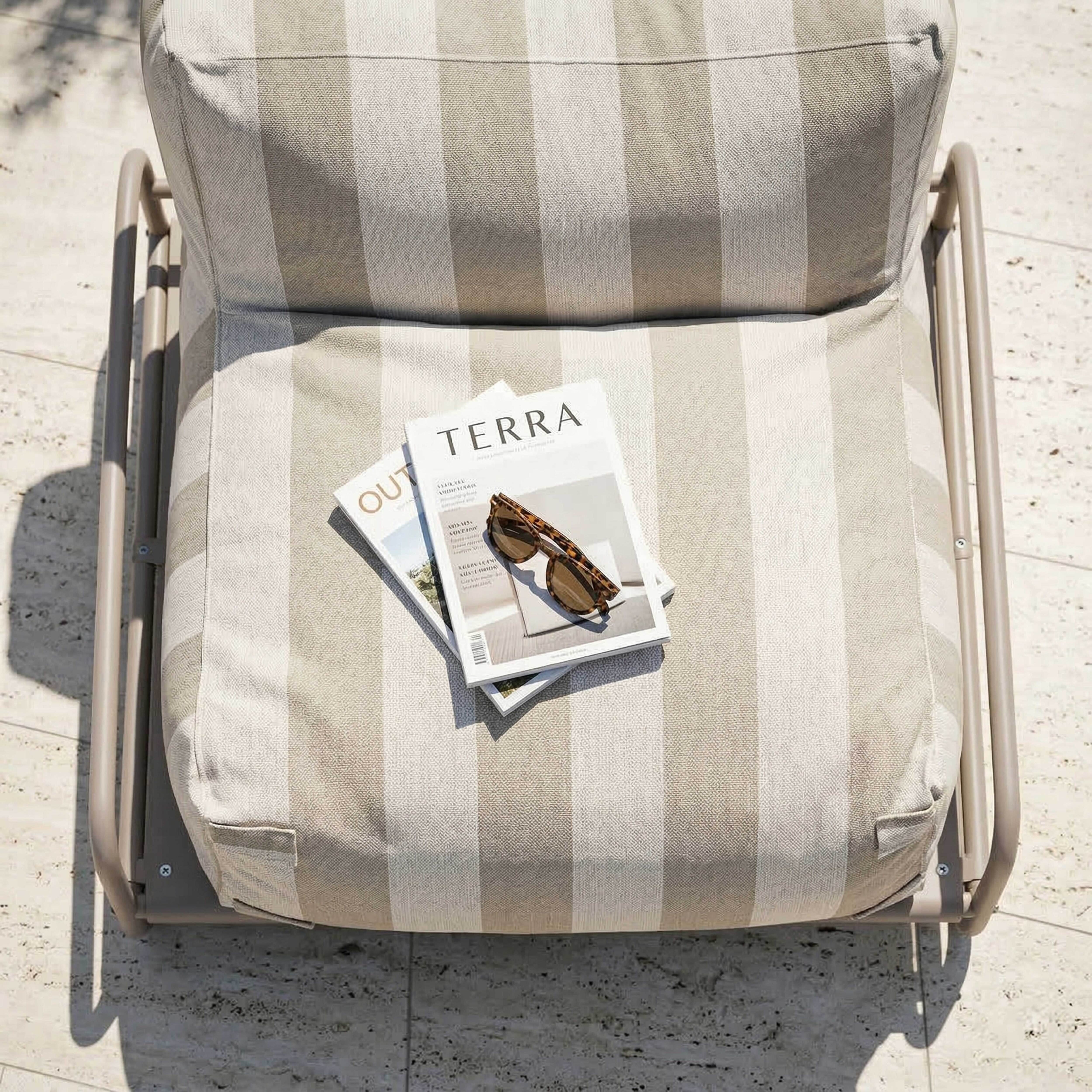 Ein gestreifter Sessel mit zwei Magazinen und Sonnenbrille auf Steinpflaster.