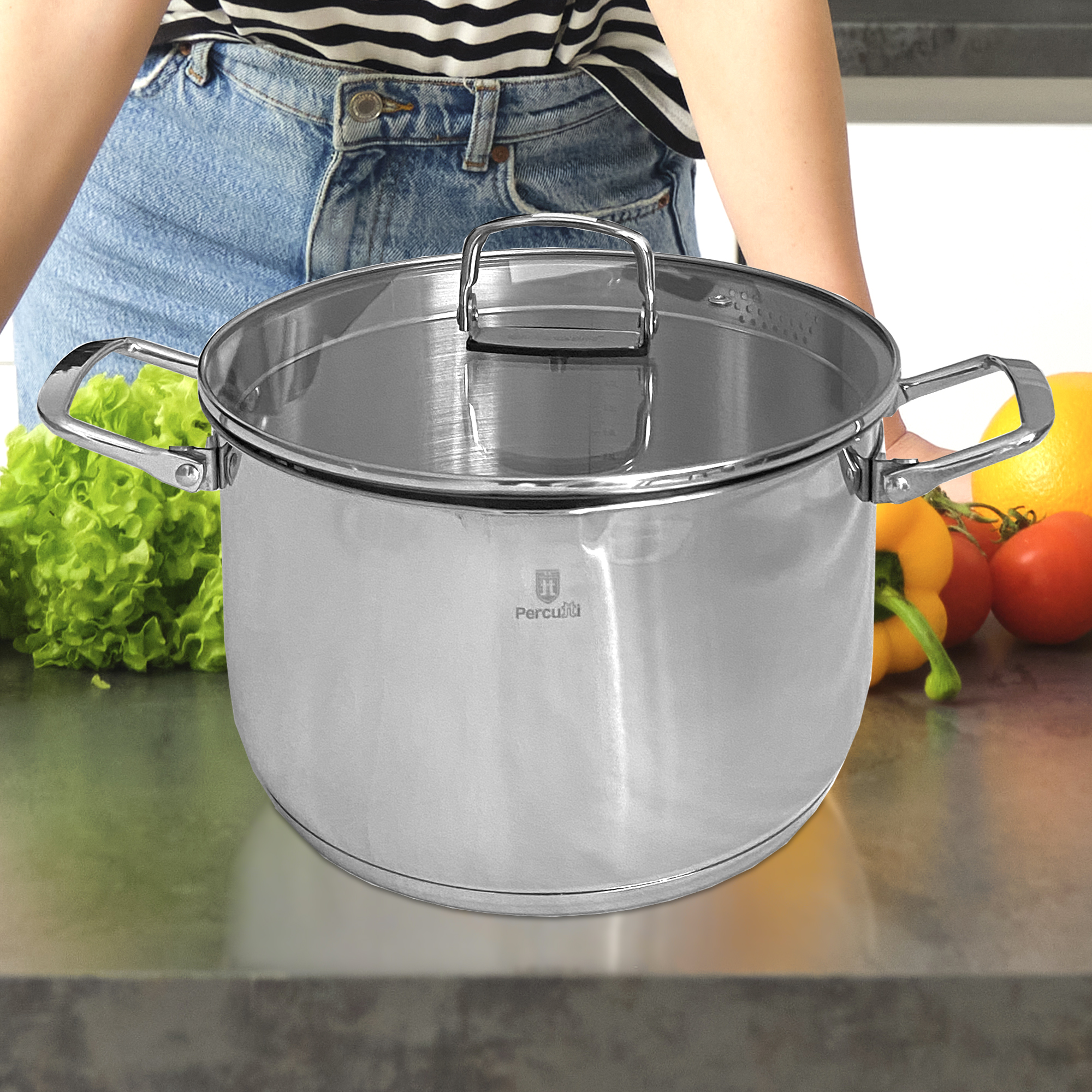 Una olla plateada con tapa de cristal está sobre una encimera junto a lechuga, pimientos, tomates y un limón.