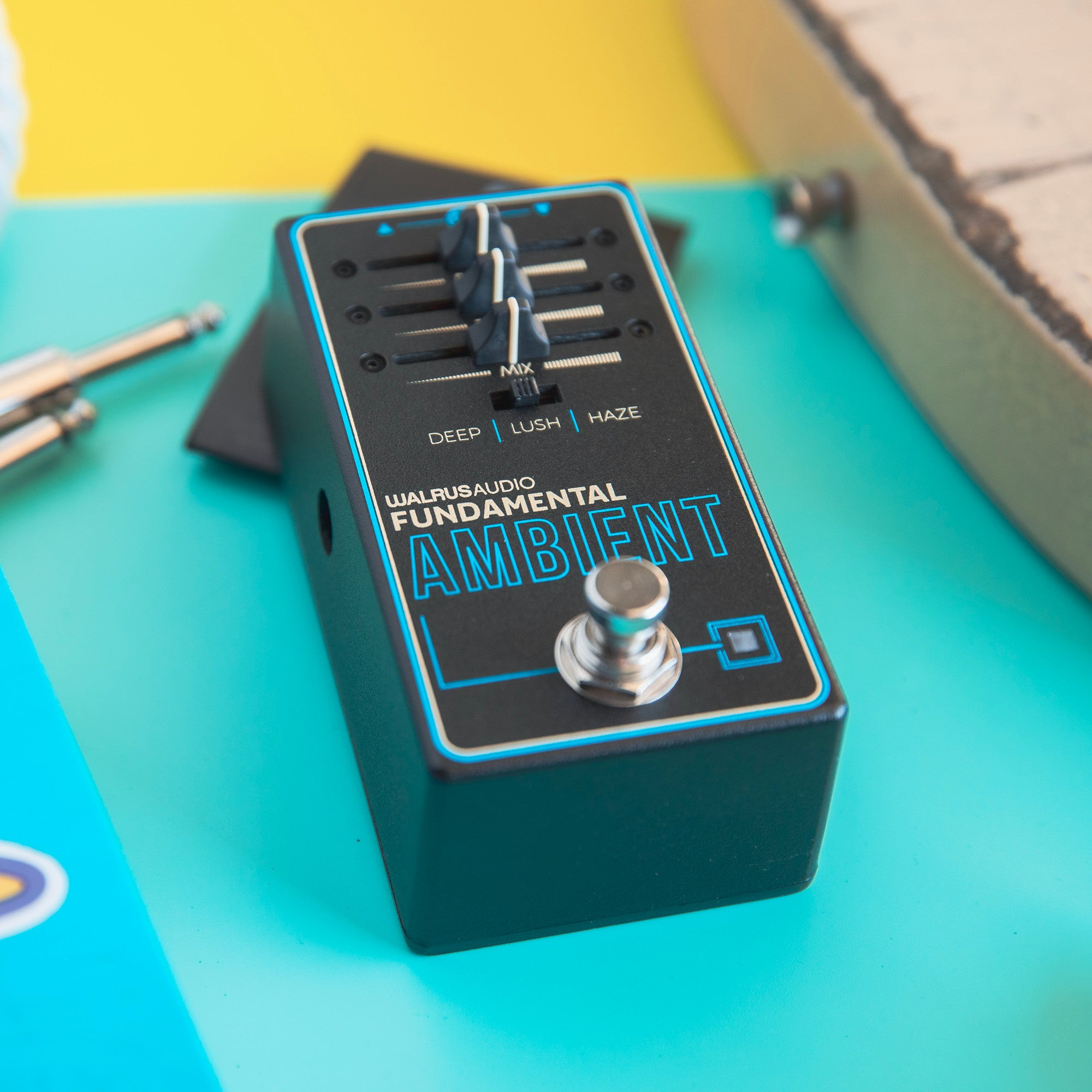 Een zwart Walrus Audio Fundamental Ambient pedaal met knoppen en voetschakelaar op een teal ondergrond.