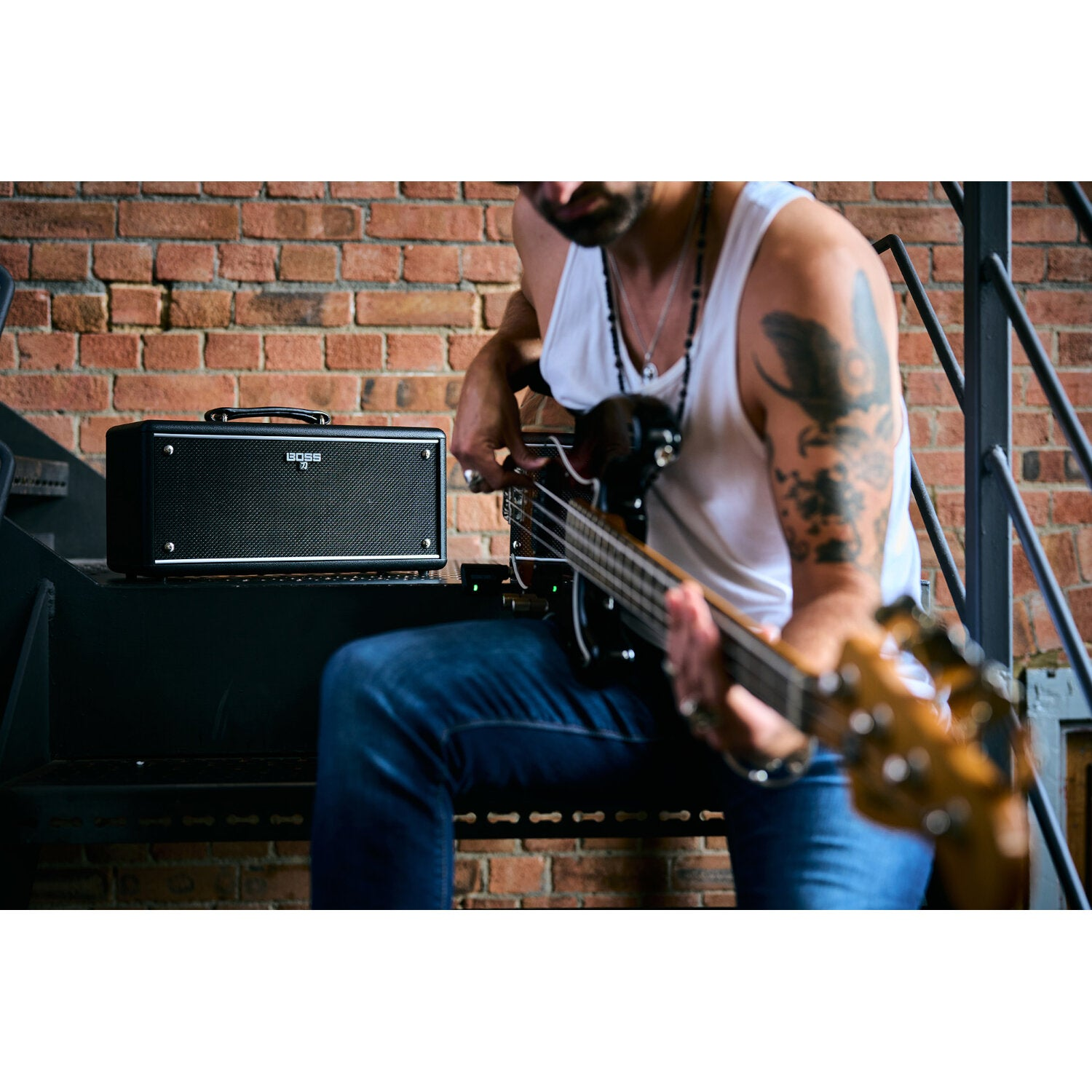 Een man in een wit hemd en spijkerbroek speelt basgitaar naast een zwarte BOSS-versterker op een metalen trap.